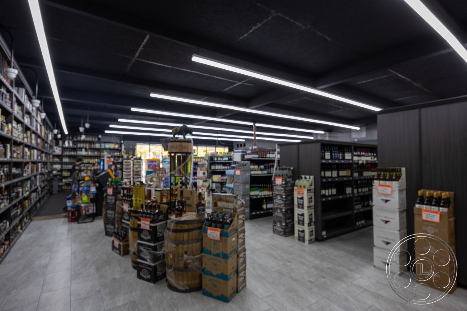 Shop 127 - open floor plan, track lighting fixtures, gray ceramic tile flooring, interior liquor store, black exposed ceiling panels, dark wooden shelving units, countertop display stands, barrel wood display tables, bottled beverage packaging, modern retail layout, contemporary retail design, spacious shopping area, clear glass window displays, vertical product arrangement