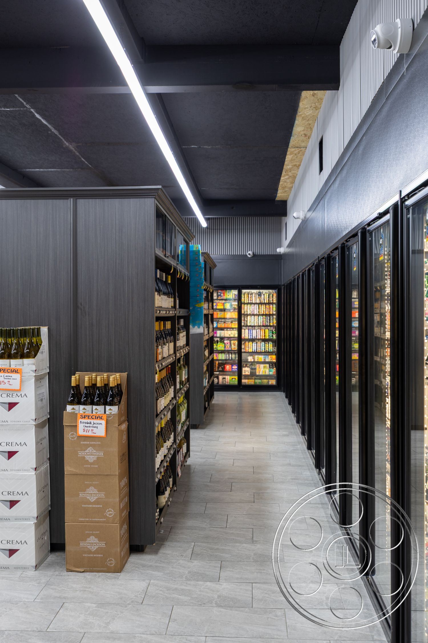 Shop 127 - high-contrast color scheme, interior shelving units with dark wood laminate finish, glossy tile flooring in light gray, exposed steel ceiling beams, fluorescent strip lighting, glass display refrigerator doors, open aisle layout for retail space, stacked wine boxes for storage, modern retail merchandise display, metal accent wall panels, bright product labels and signage, minimalistic product organization, spacious corridor design, efficient aisle navigation, ample natural lighting from large windows