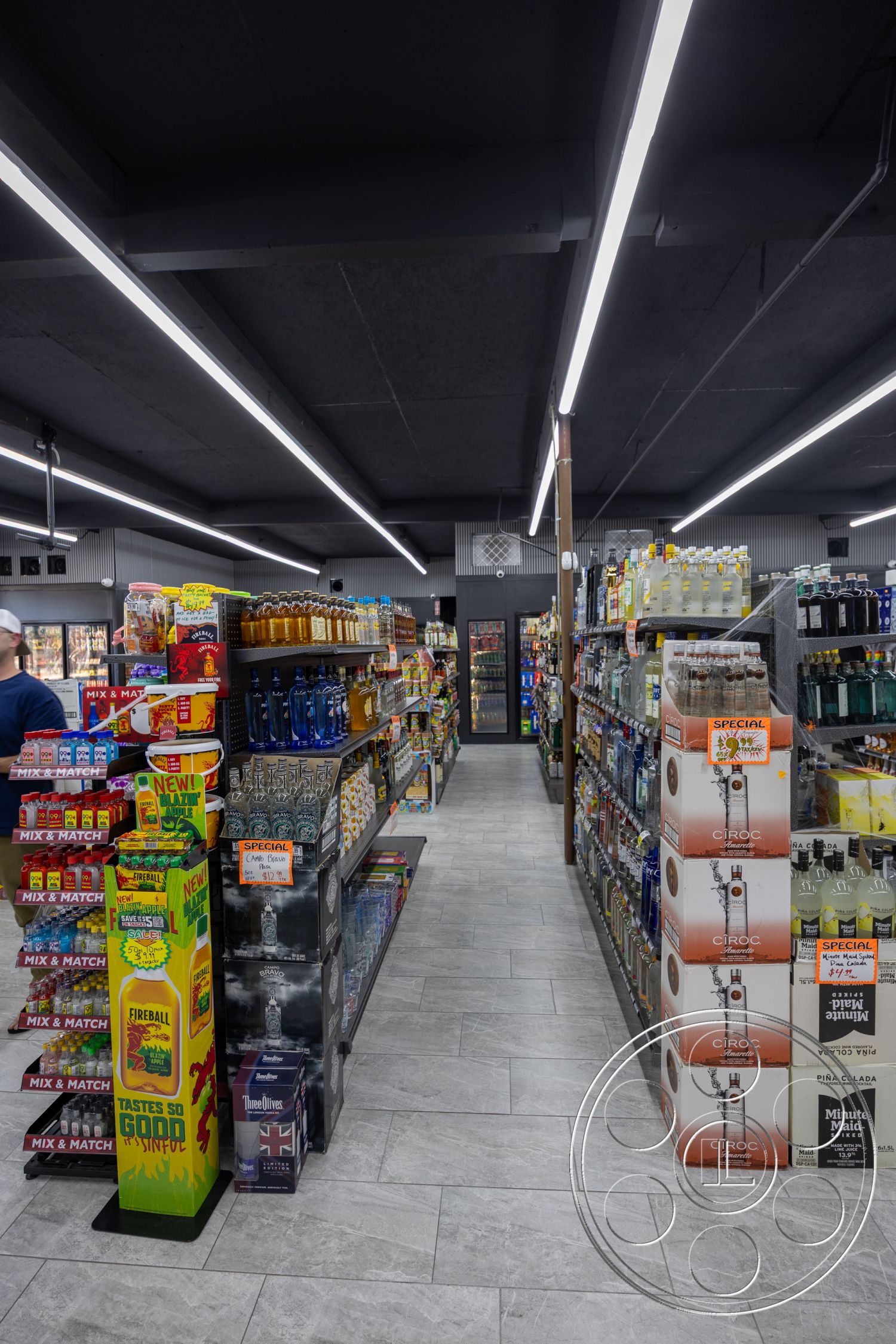 Shop 127 - open floor plan, ambient lighting, metal shelving units, grey porcelain tile flooring, LED strip lighting, modern retail design, interior store layout, high ceiling with exposed beams, product display stands, refrigerated drink aisle, parallel aisle configuration, merchandising signage, bottled beverage displays, contemporary store fixtures, counter space for checkout