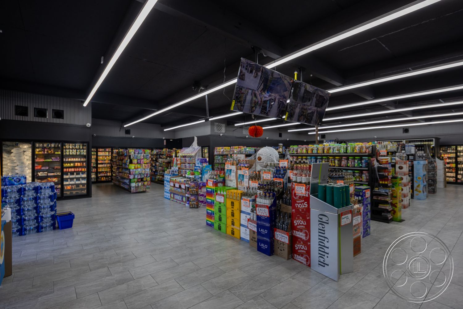 Shop 127 - open floor plan, concrete block walls, gray tile flooring, stacked cardboard boxes, industrial shelving units, interior grocery store, warehouse-style interior, colorful product packaging, modern retail layout, suspended LED lighting, black drop ceiling, glass refrigerator units, metal display shelves, LED illuminated signage, refrigerated drink section, aisle displays, ambient retail lighting, wall-mounted televisions, consumer goods display, low-profile merchandise