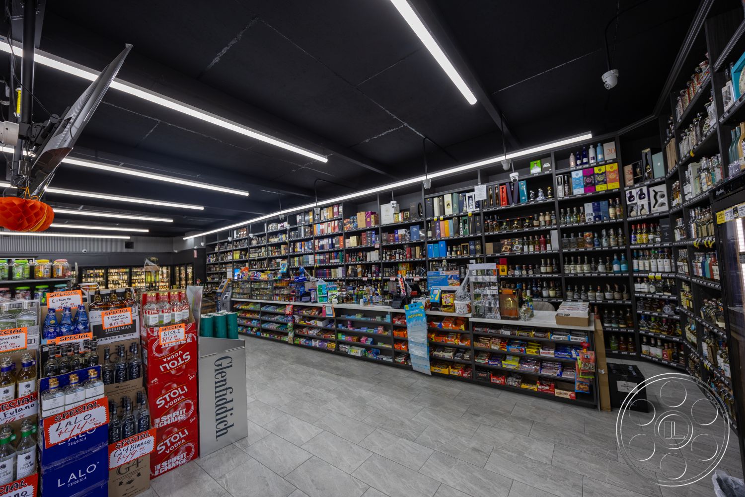 Shop 127 - glass display cases, concrete tile flooring, colorful product packaging, refrigerated beverage coolers, cash register area, interior liquor store layout, black drop ceiling tiles, LED strip lighting installation, retail shelving units, wall-mounted product displays, white accent wall, industrial-style shelving, merchandise end caps, open floor plan shopping space, organized product arrangement, modern retail fixtures, accessible store aisles, customer service counter, themed promotional displays, store signage fixtures