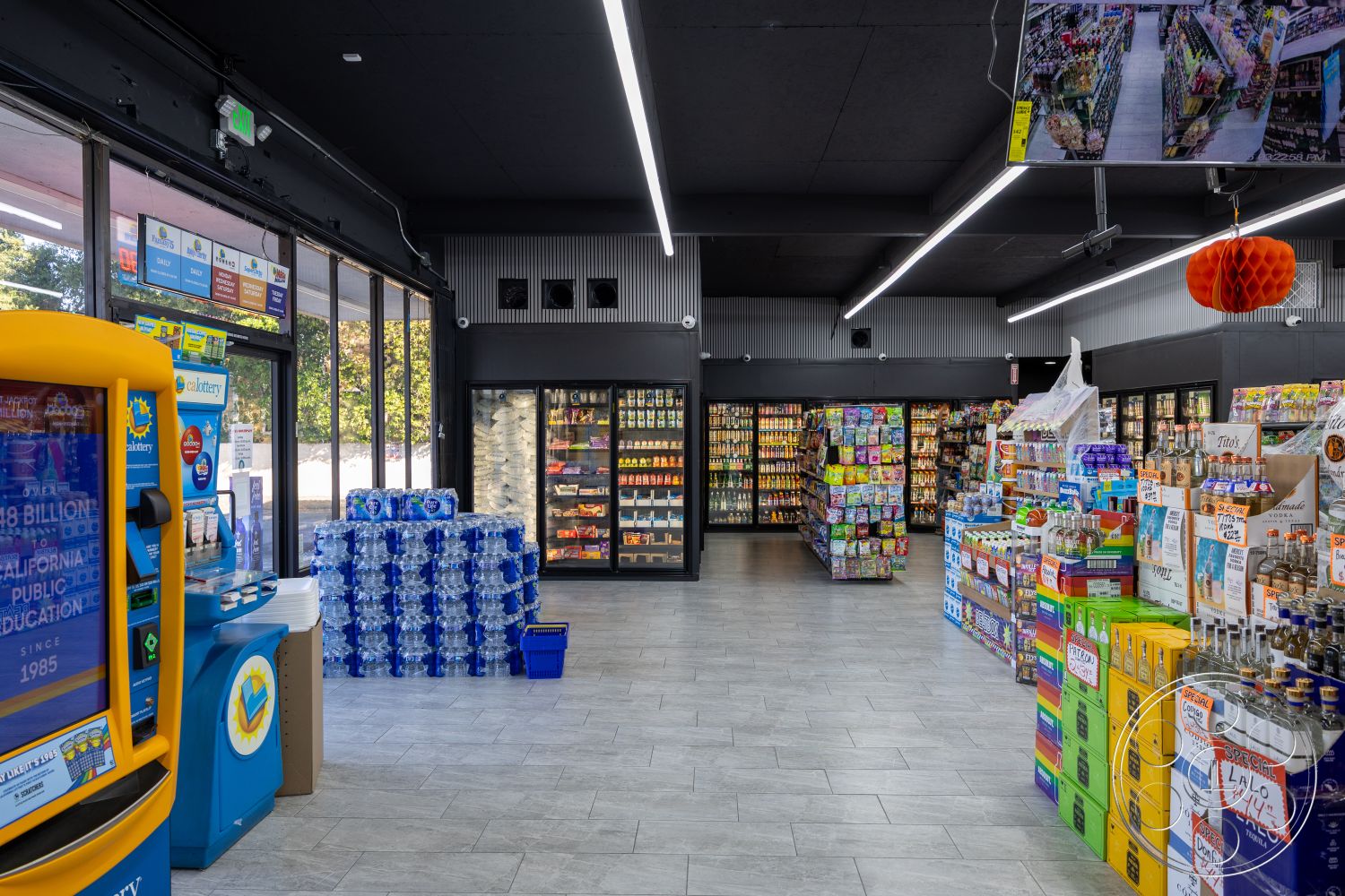 Shop 127 - open floor plan, metal shelving units, polished tile flooring, black suspended ceiling panels, large glass storefront windows, interior grocery store, brightly lit shopping environment, colorful product packaging, fluorescent lighting strips, merchandise display racks, water bottle stack display, refrigerated food display cases, blue plastic bins, lottery ticket kiosk, urban convenience store setting