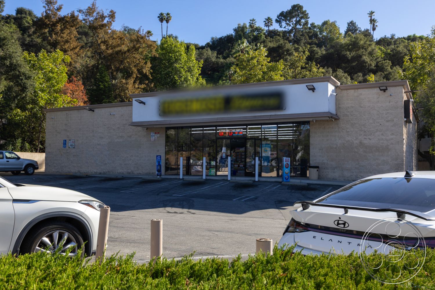 Shop 127 - green landscaping, metal light fixtures, asphalt parking lot, commercial retail space, large glass storefront windows, exterior stone facade, urban convenience store, flat roof commercial building, screened sidewalk entry, paved parking spaces, bushes along facade, surrounding hillside setting