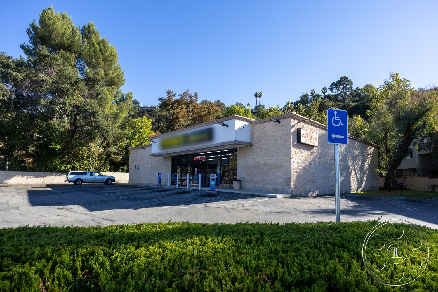 Shop 127 - exterior, landscaped greenery, stucco facade, spacious outdoor area, surrounded by trees, vibrant urban setting, single-story building, concrete parking lot, flat roof design, urban commercial space, large glass storefront windows, handicap accessible parking, decorative signage, naturally shaded environment, concrete approach, visible pedestrian accessibility