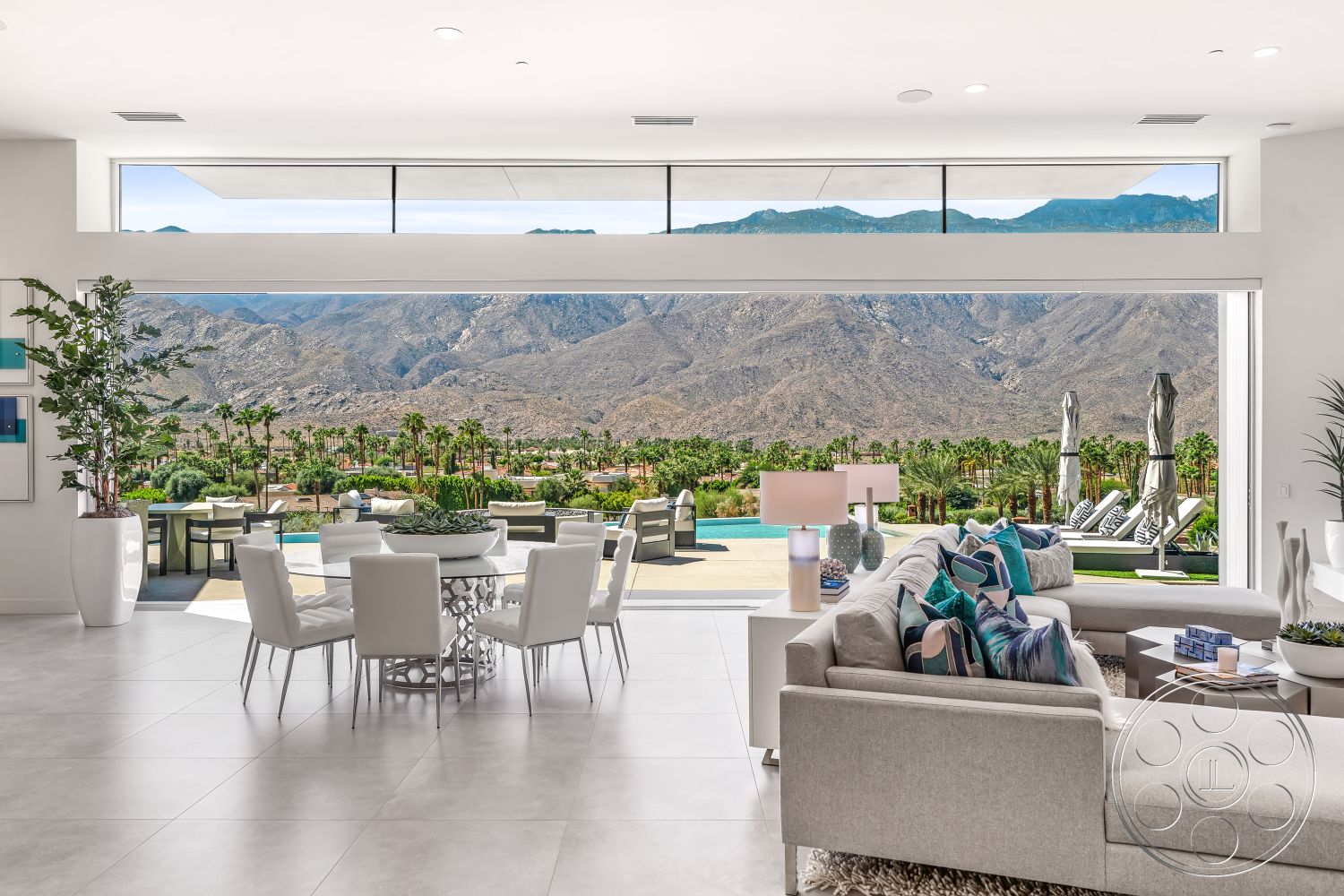 Palm Springs 109 - large glass sliding doors, open floor plan, large windows for natural light, mountain backdrop, bright and airy atmosphere, colorful accent pillows, modern table lamp, stone landscaping, poolside lounging area, circular dining table, minimalist furniture design, potted indoor plants, contemporary sectional sofa, concrete patio floor, interior modern living room, white upholstered dining chairs, rectangular swimming pool view, light grey tile flooring, palm trees in exterior setting, urban desert environment, view of outdoor seating