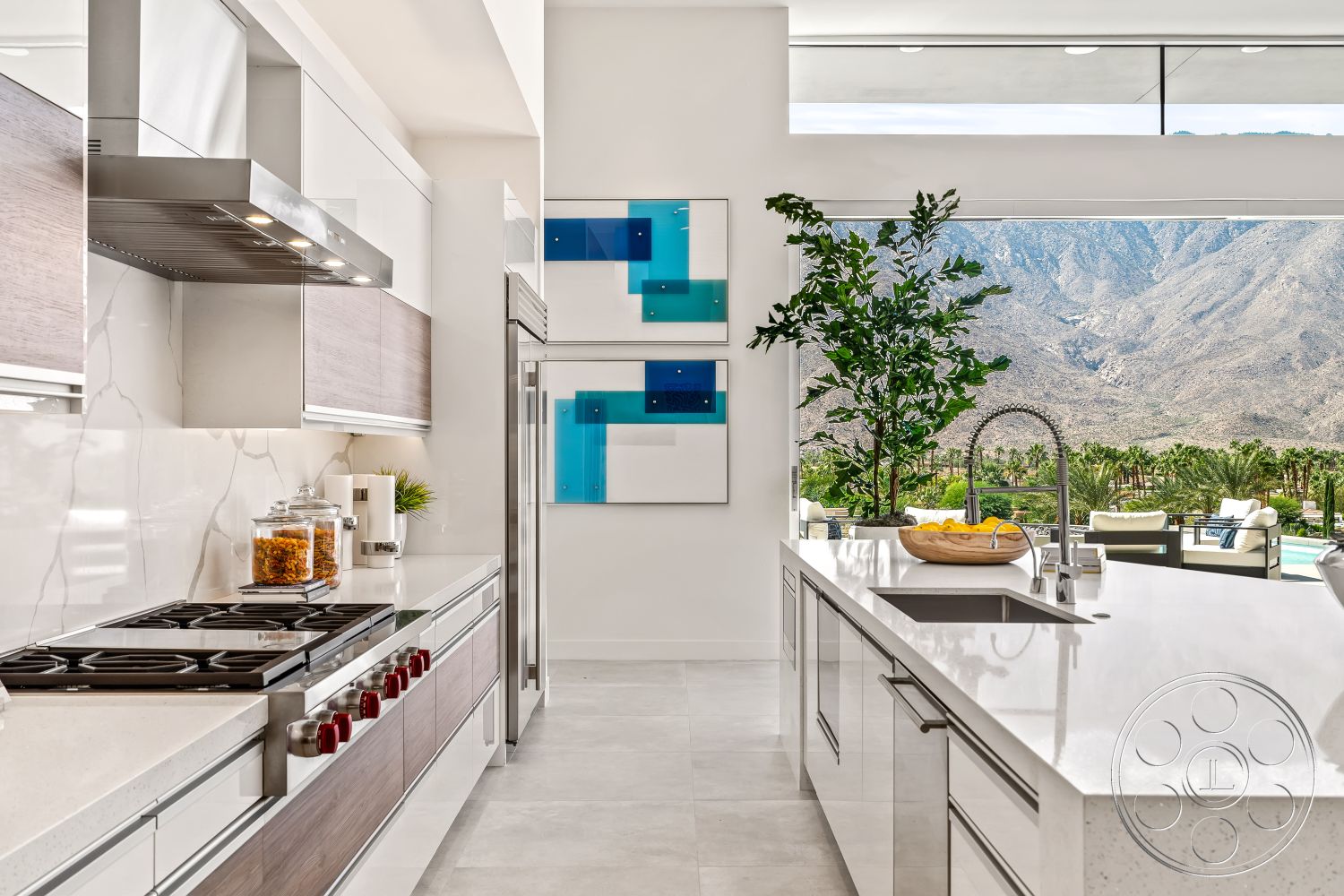 Palm Springs 109 - modern kitchen faucet, stainless steel range hood, minimalist kitchen design, interior kitchen with quartz countertops, modern cabinetry with wood veneer finish, marble backsplash with veining details, open concept layout with floor-to-ceiling windows, contemporary decorations with abstract art, white tile flooring, kitchen island with integrated sink, indoor greenery with potted plant, outdoor mountain view, high-end appliances including built-in refrigerator, dual-tone cabinetry featuring light and dark wood, bright and airy kitchen space, luxurious kitchen with recessed lighting