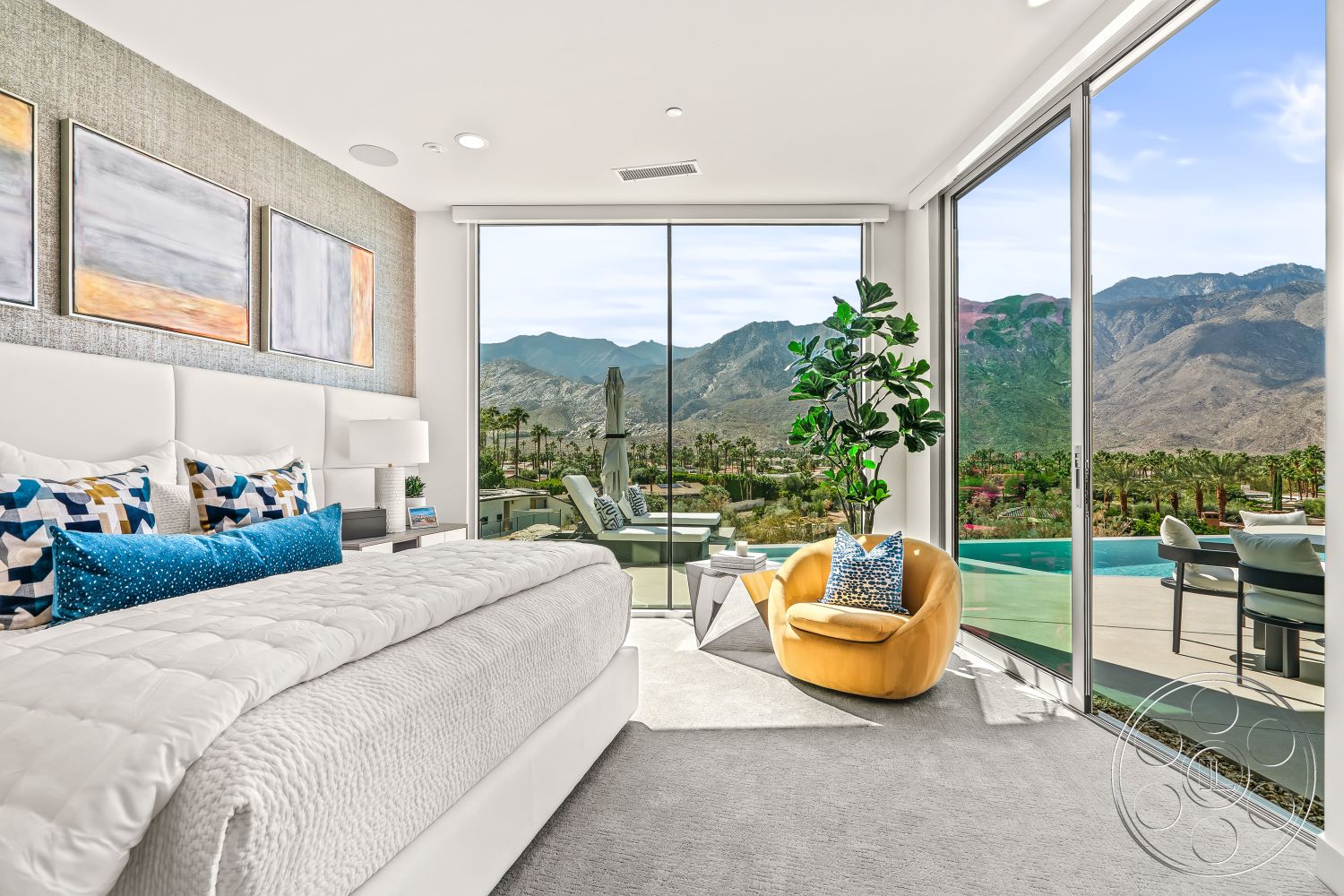 Palm Springs 109 - indoor potted plant, exposed stone accent wall, king-size bed with white bedding, large floor-to-ceiling glass windows, interior bedroom with white upholstered headboard, plush carpet flooring in neutral grey, decorative throw pillows in blue and geometric patterns, mountain view from the bedroom, sunny modern interior design, metallic geometric side table, stylish yellow accent chair with blue pillows, minimalist artwork with soft tones, open layout connecting to outdoor patio, outdoor swimming pool view, contemporary architecture style