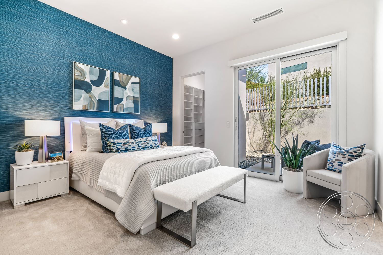 Palm Springs 109 - neutral color palette, open floor plan, bright natural light, glass sliding door, interior bedroom, contemporary furniture design, contemporary art wall decor, light gray carpet flooring, minimalist bedside lamps, textured blue accent wall, white upholstered bed, decorative throw pillows with blue patterns, modern nightstands, plant in white ceramic pot, white room trim, built-in closet shelving
