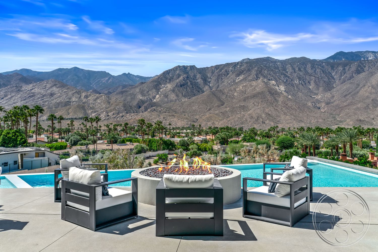 Palm Springs 109 - minimalist design elements, modern architectural style, open-air terrace, drought-tolerant landscaping, desert environment, contemporary outdoor living space, exterior patio with concrete flooring, rectangular infinity pool with glass tiles, modern fire pit with lava stones, aluminum outdoor seating with white cushions, mountain view backdrop with palm trees, landscaped desert garden, sunny poolside area, sleek glass railing, luxury outdoor escape