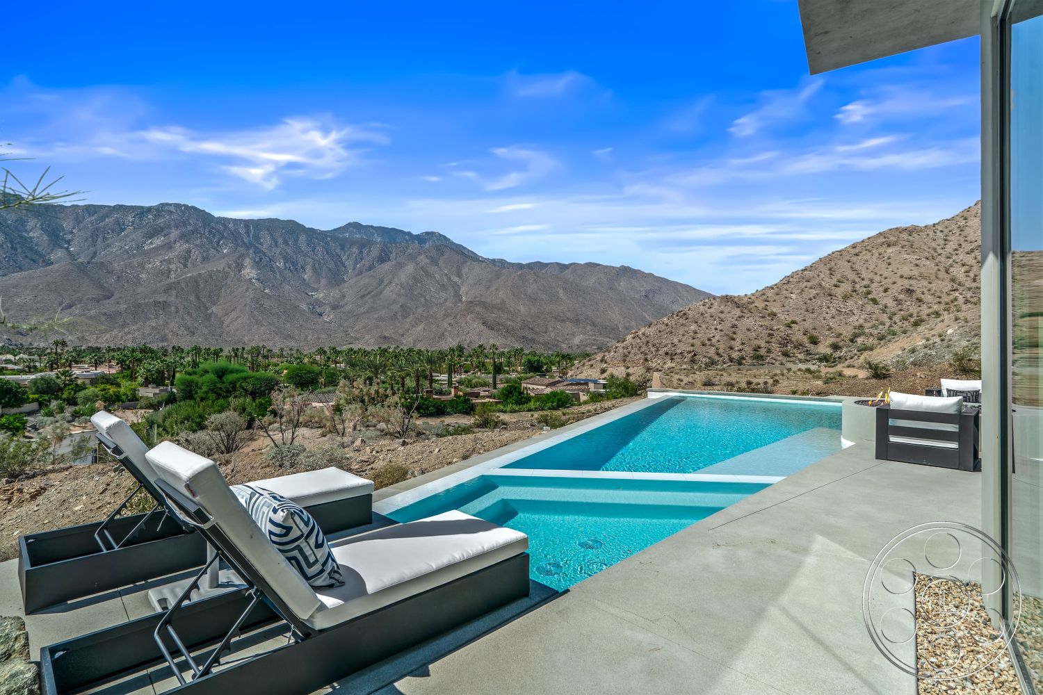 Palm Springs 109 - modern lounge chairs, mountain view setting, floor-to-ceiling glass windows, concrete pool border, minimalist architectural style, exterior concrete patio flooring, open-air pool area, infinity pool with rectangular shape, desert home exterior, landscaped yard with desert plants, serene natural landscape, contemporary outdoor lounging area, clear blue skies, textured concrete steps leading to pool
