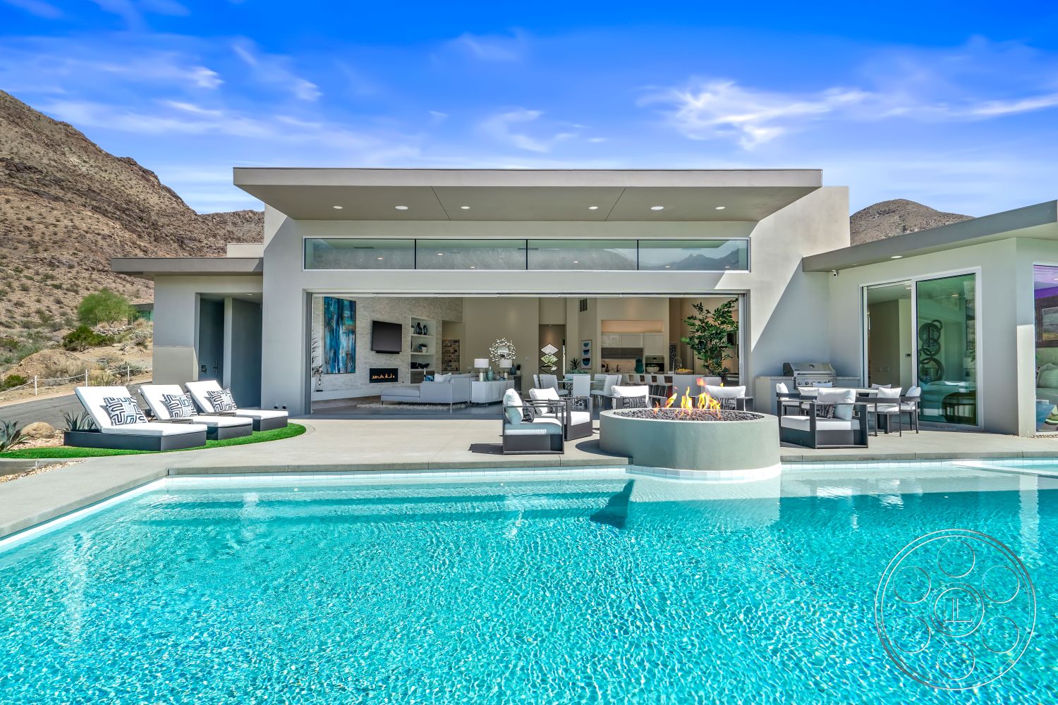 Palm Springs 109 - white stucco walls, large glass sliding doors, modern aesthetic, rectangular swimming pool, landscaped garden area, open-concept living area, interior white painted walls, fire pit with stone surround, minimalist outdoor furniture, exterior concrete pool deck, gray concrete pavers, horizontal window design, flat-roofed modern architecture, mountainous desert backdrop, gathering space by the pool, sleek lines and open spaces