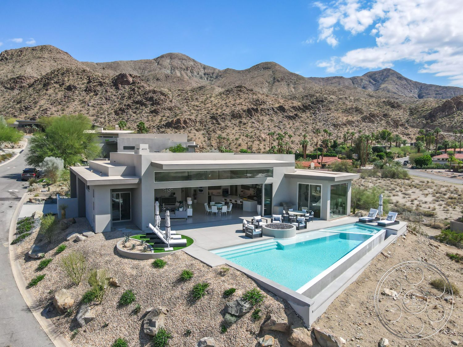 Palm Springs 109 - natural stone accents, modern outdoor furniture, floor-to-ceiling windows, outdoor dining area, circular driveway, open-concept living space, infinity pool, glass fire pit, concrete pool deck, rectangular pool, minimalist exterior, sunny exterior, stucco exterior walls, concrete patio floor, modern architectural design, desert home setting, landscaped rock garden, wood seating area, surrounding mountain view, desert backdrop, desert landscaping elements
