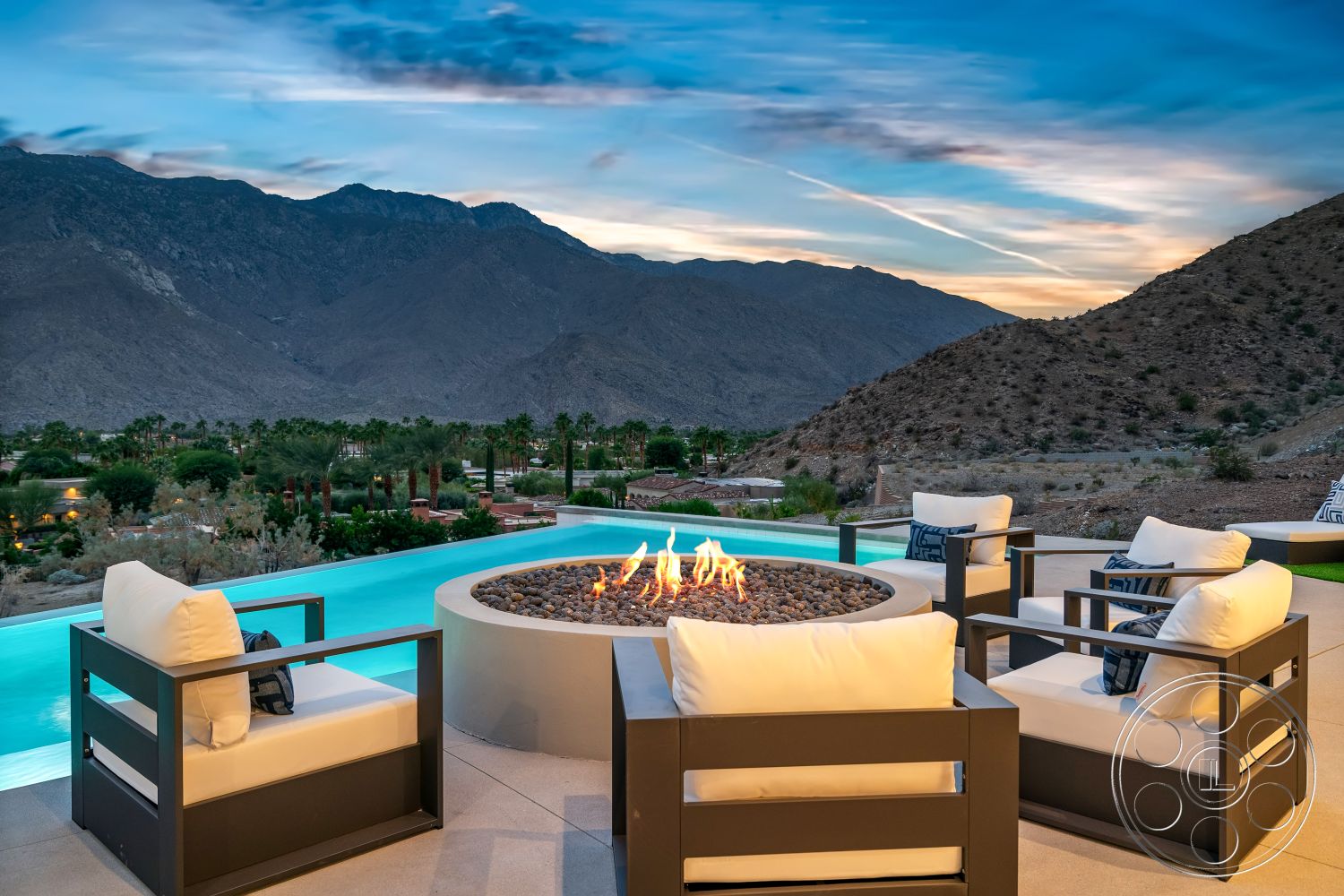 Palm Springs 109 - interior-exterior flow, desert landscape, outdoor lounging area, rectangular pool, glass panel fencing, mountain view setting, landscaped garden area, concrete patio flooring, textured concrete surfaces, infinity pool with concrete coping, fire pit with decorative stones, modern outdoor seating chairs, slip-resistant tile surrounding pool, evening ambiance with sunset, fire pit centerpiece