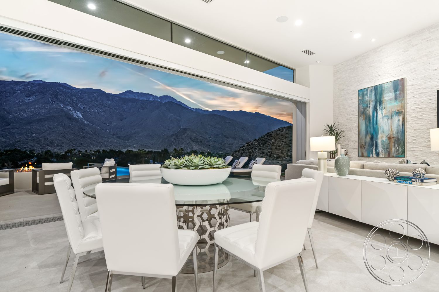 Palm Springs 109 - open concept living space, bright and airy atmosphere, contemporary design aesthetic, textured stone accent wall, light gray tile flooring, floor-to-ceiling windows with mountain view, contemporary artwork on wall, interior dining area with glass table, modern white leather dining chairs, indoor-outdoor flow with large sliding doors, minimalist white cabinetry, succulent centerpiece on table, mountain landscape backdrop, elegant linear lighting fixtures, seating area with modern gray sofa
