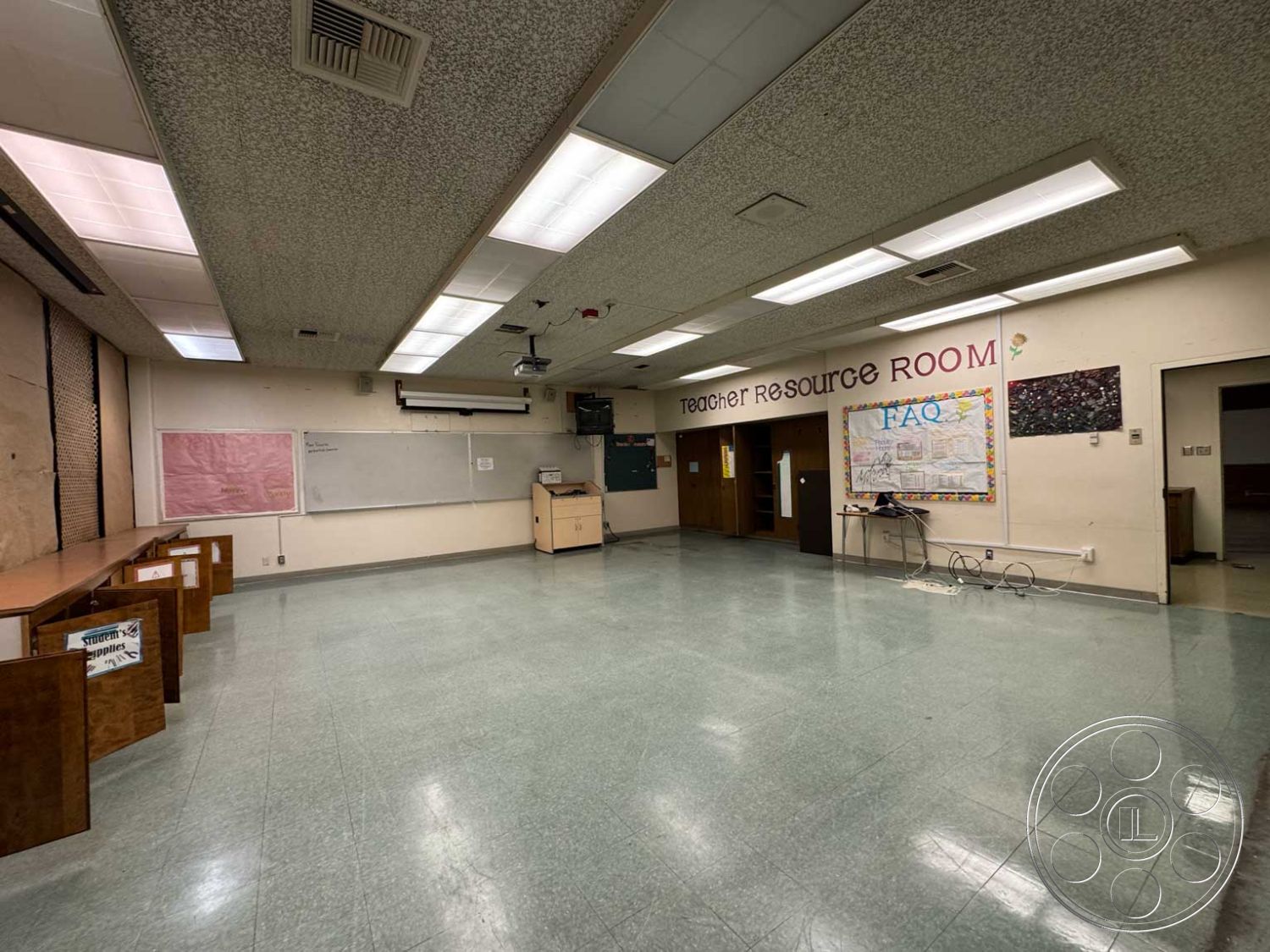 School 3 - neutral wall color, textured plaster walls, vinyl flooring, chalkboard wall, acoustic ceiling panels, fluorescent overhead lighting, interior classroom space, spacious room layout, wooden teacher resource desk, dry erase whiteboard, open doorway access, bulletin board area, cabinet storage units