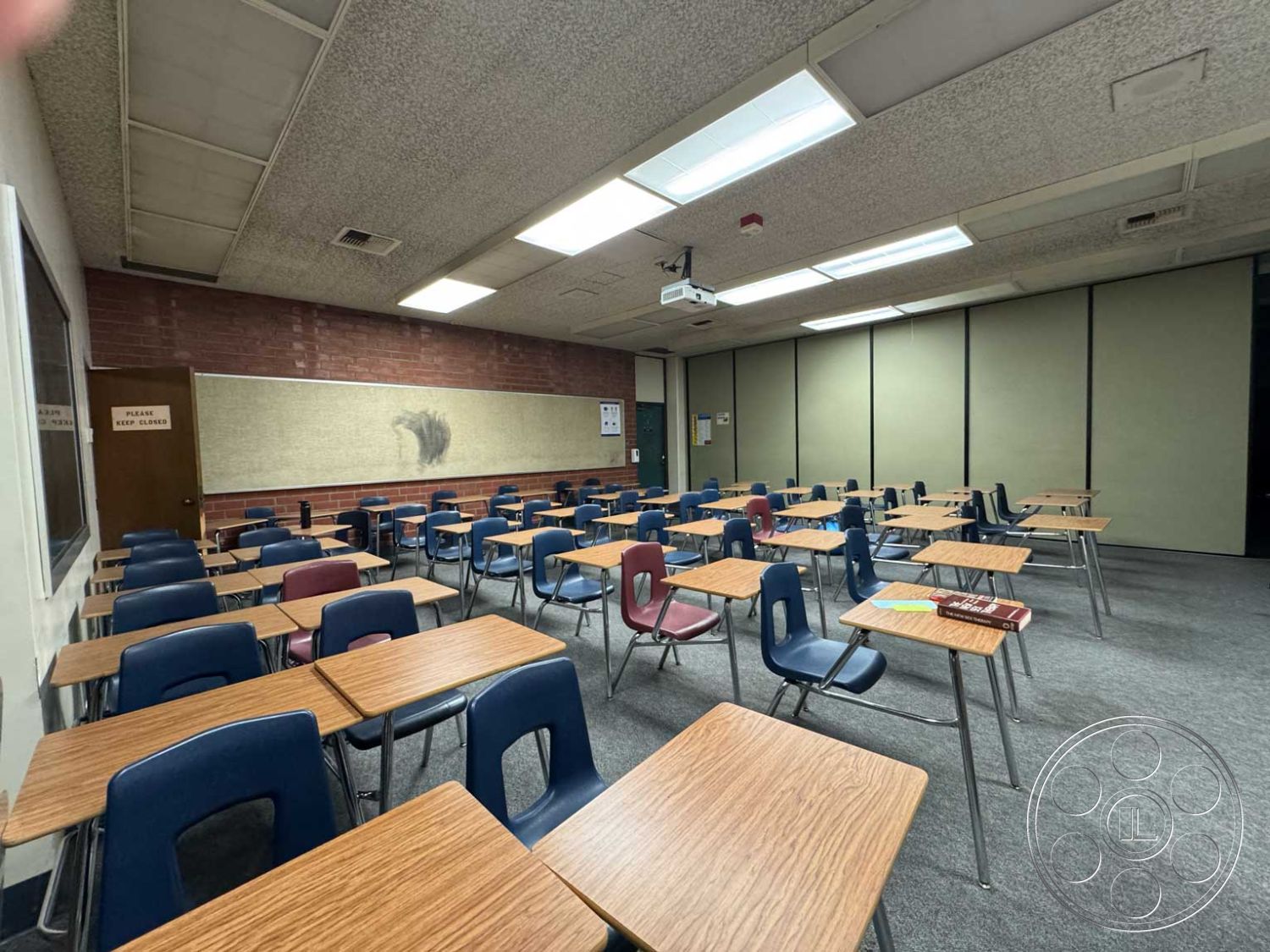 School 3 - overhead fluorescent lighting, exposed brick accent wall, acoustic tile ceiling, interior classroom setting, projector mounted on ceiling, faux wood laminate flooring, steel-framed classroom desks, blue plastic stacking chairs, neutral beige wall panels, standard classroom layout, wooden door entrance, large wall-mounted bulletin board