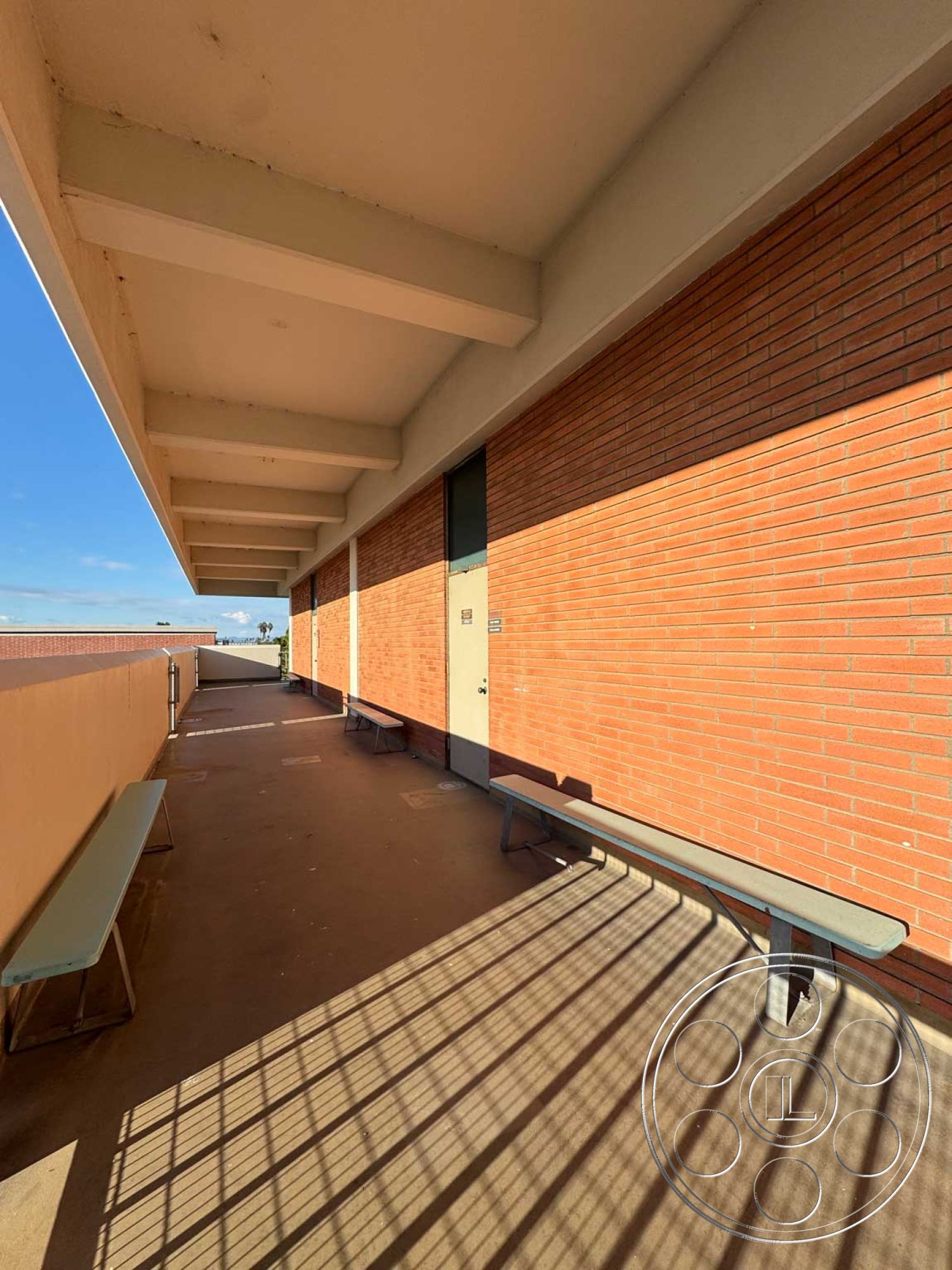 School 3 - urban setting, concrete walkway, minimalist architectural style, exterior brick wall, flat concrete surface, sunlit exterior space, open-air corridor, modern bench seating, horizontal concrete ceiling beams, rectangular door, textured brick finish, shadow patterns on floor