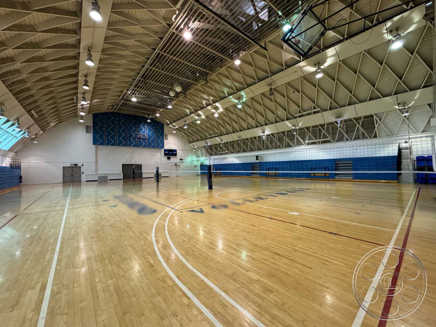 School 3 - industrial lighting fixtures, open floor plan design, hardwood gymnasium flooring, exposed steel roof trusses, vaulted ceiling with skylights, blue acoustic wall panels, court markings for volleyball, interior sports facility, multi-purpose activity space, green wall accents, seating benches along walls, high ceiling with angled design, glass viewing windows