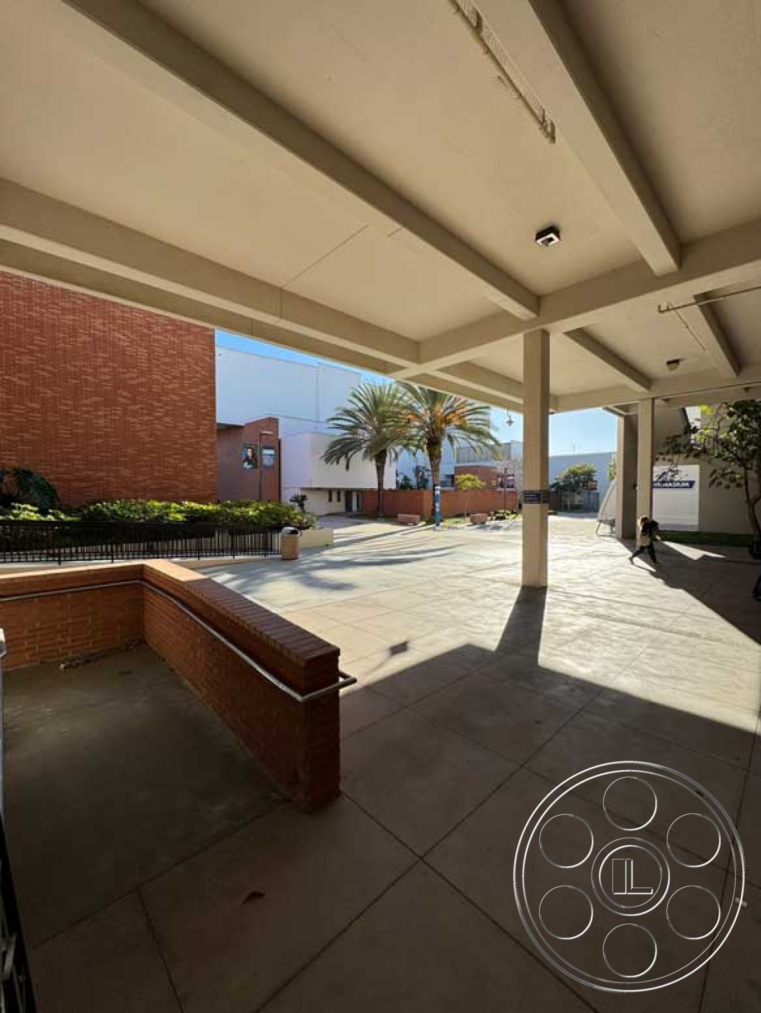 School 3 - surrounding commercial buildings, large outdoor space, modern architectural details, open courtyard design, textured concrete patio flooring, exposed concrete ceiling beams, brick planter wall, urban exterior setting, lush landscaping with palm trees, cityscape view from patio