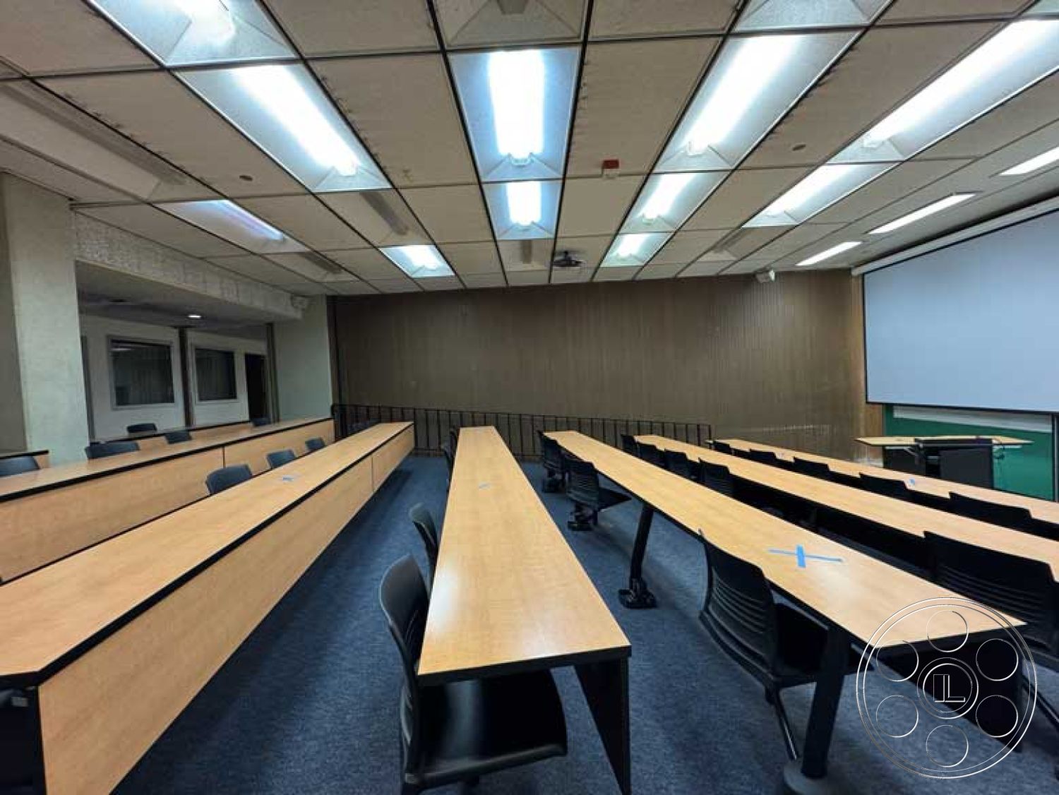 School 3 - carpet flooring, acoustic ceiling tiles, wood panel accent wall, fluorescent overhead lighting, interior classroom, projector screen wall, laminate conference tables, black plastic seating, media presentation area, linear room layout, multi-purpose learning space, row seating arrangement