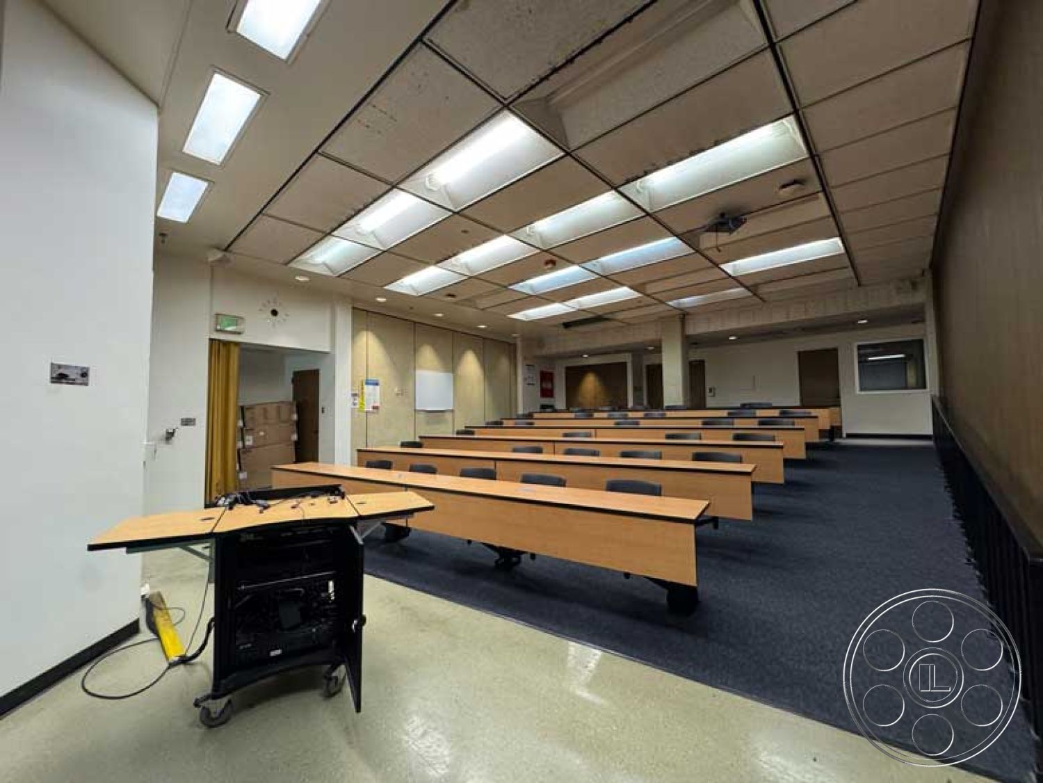 School 3 - white walls, interior space, vinyl flooring, fiberboard drop ceiling tiles, flourescent light fixtures, wood laminate classroom tables, plastic folding chairs, built-in projection screen, grey accent wall, classroom layout, educational room, university classroom, exposed ceiling infrastructure, divider wall, temporary storage area, rollaway teaching cart