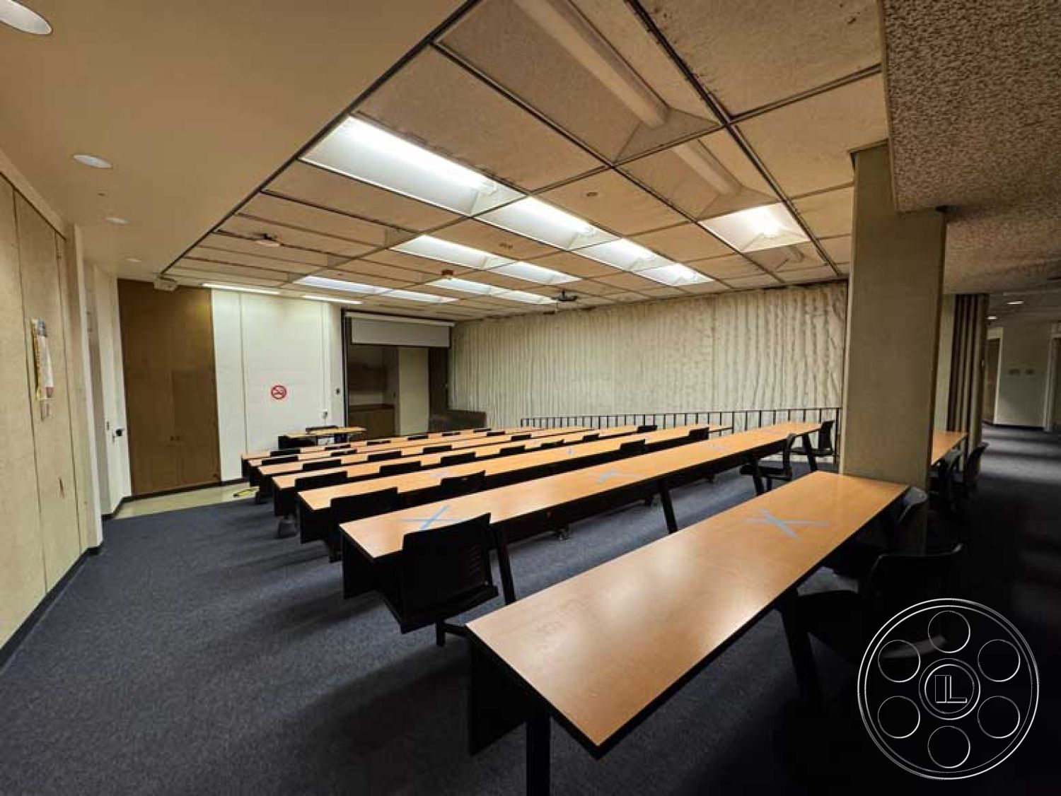 School 3 - natural light from windows, black plastic chairs, acoustic ceiling tiles, plush carpet flooring, interior classroom design, drywall partition walls, wooden lecture tables, rear projection screen setup, linear fluorescent lighting, closed door access, rows of classroom seating, simple contemporary style, functional educational space, multi-purpose room setting