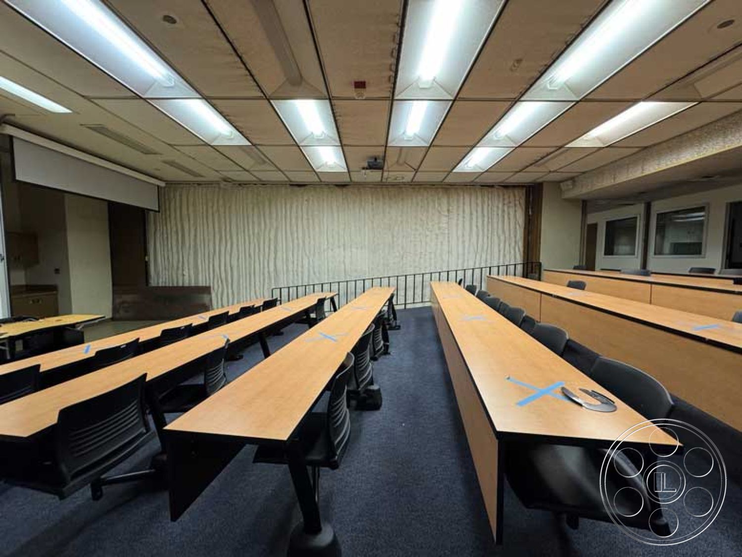 School 3 - carpeted flooring, acoustic panel walls, black ergonomic office chairs, textured plaster wall accents, drop ceiling with fluorescent lighting, modern educational environment, interior classroom design, wooden classroom tables, large projector screen mounting, open lecture space layout, straight row seating arrangement, metal railing at the front