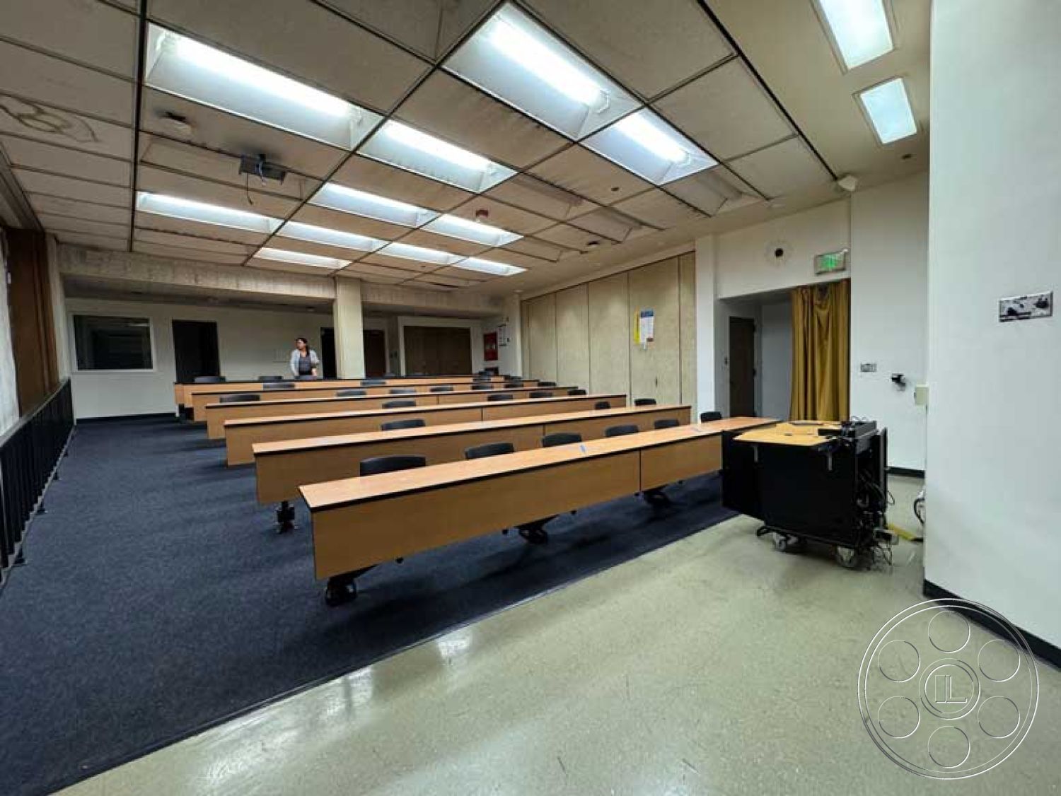 School 3 - minimalist design elements, industrial lighting fixtures, multi-purpose space, double door entry, blackboard wall, white painted walls, fluorescent ceiling lights, light acoustic ceiling tiles, interior classroom, rectangular classroom layout, carpet floor tiles, wooden student desks, plastic classroom chairs, projector stand, curtained side entrance, lecture hall setting