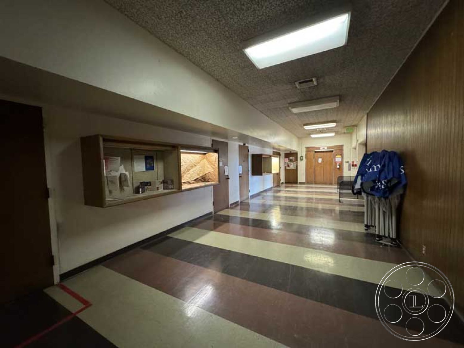 School 3 - textured wall surfaces, brown wood paneling, interior hallway, acoustic tile ceiling, fluorescent ceiling lights, functional circulation space, laminate flooring with striped pattern, glass display case with wooden frame, metal coat rack, painted drywall, internal doorways with wood doors, ample natural light from ceiling fixtures, buildings with institutional design elements