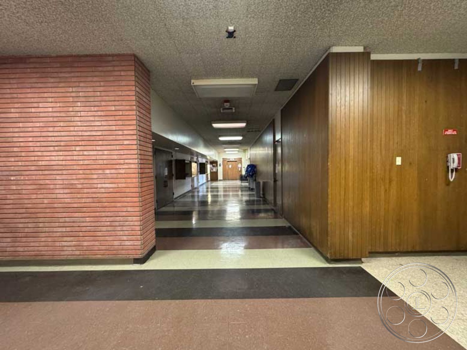 School 3 - exposed brick accent wall, wood paneled walls, commercial building interior, functional corridor layout, vinyl tile flooring, black and white checkered floors, linear hallway design, modern fluorescent lighting fixtures, double doors at the end of the hall