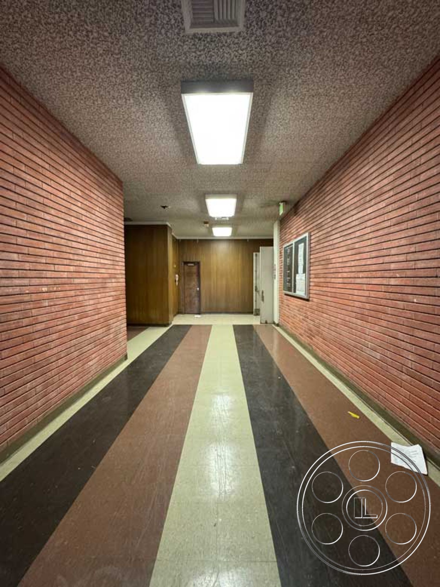 School 3 - commercial building interior, exposed brick interior wall, vinyl tile flooring, drop ceiling with fluorescent lights, modern geometric shapes, glass notice board, striped patterned floor, wood paneling interior door, corridor hallway
