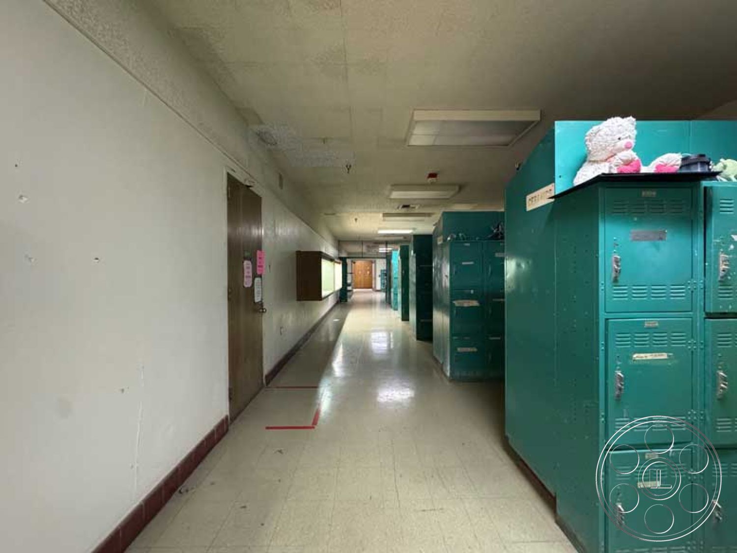 School 3 - school environment, interior hallway, painted drywall walls, drop ceiling panels, vinyl tile flooring, wide corridor layout, linear fluorescent lighting, linear hallway design, steel storage lockers, wooden classroom doors, colorful locker accents, institutional lighting fixtures, adjacent classroom access, utility wall-mounted cabinetry, functional space for education