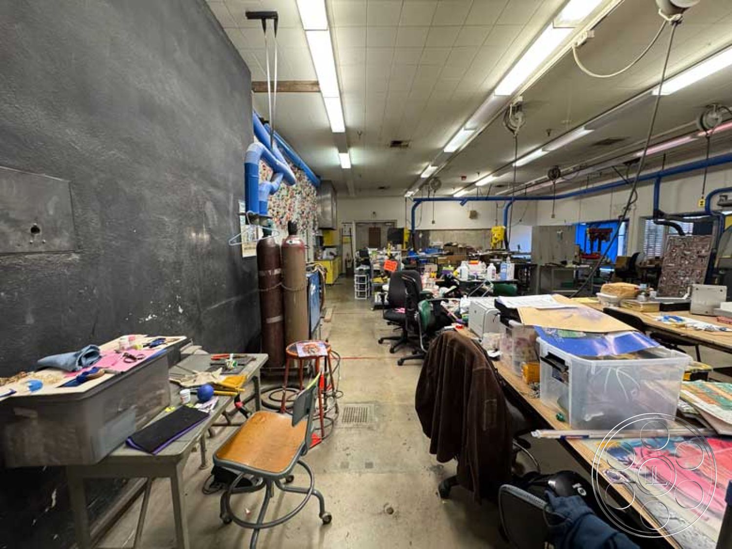 School 3 - concrete floor, task lighting, creative studio space, industrial ceiling tiles, interior workspace, rolling office chairs, wooden work tables, exposed metal pipes, paint splatters on surfaces, black chalkboard wall, mixed-use workshop environment, metalwork stations, storage bins and containers, varied crafting materials, colorful fabric remnants, sewing machines setup, workshop tools, multi-purpose work area, scattered creative supplies