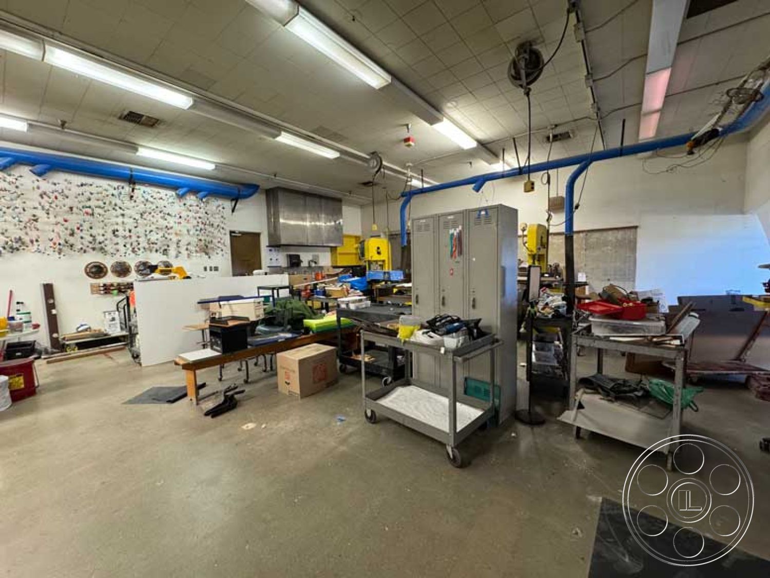 School 3 - concrete flooring, metal shelving units, exposed ductwork, stainless steel countertops, functional workshop environment, interior industrial workspace, wooden workbenches, ceilings with fluorescent lighting, blue painted pipe insulation, storage lockers, assorted tools and equipment, organized chaos workspace, multicolored tool wall installation