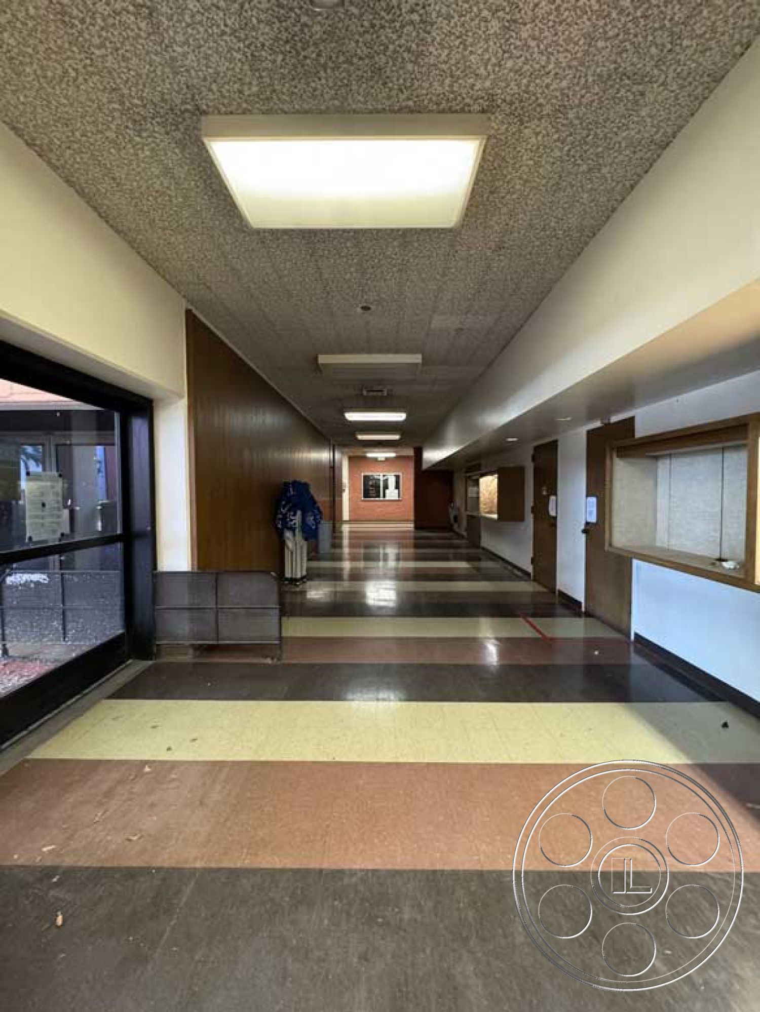 School 3 - neutral color palette, terrazzo flooring, glass entry door, natural light from windows, interior hallway, modernist architectural style, wood paneling wall, public building interior, drop ceiling with fluorescent lights, plastered wall finish, open layout corridor, mixed color floor tiles, decorative wall signage, built-in display cases, coatrack by entry