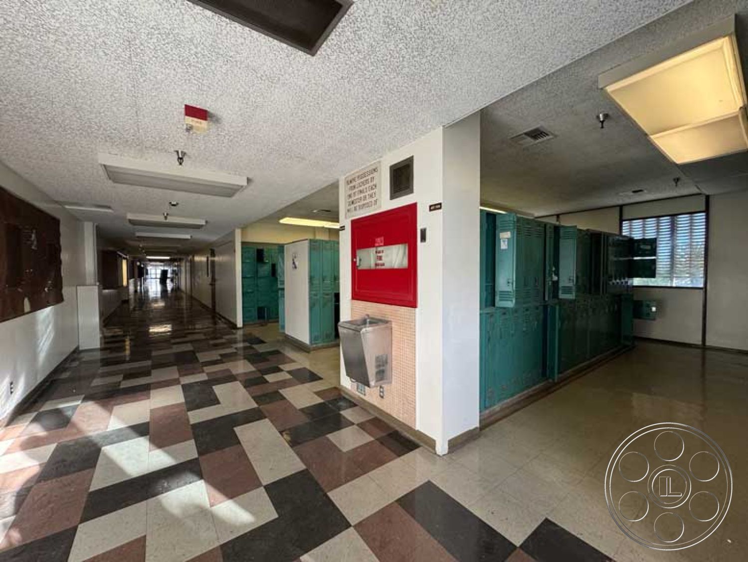 School 3 - acoustic ceiling tiles, interior hallway, horizontal window blinds, textured plaster wall panels, open corridor layout, fluorescent ceiling lights, vinyl tile flooring, checkerboard pattern floor tiles, lightweight concrete block walls, green metal lockers, red metal fire alarm box, drinking fountain, public school architectural style, educational facility design