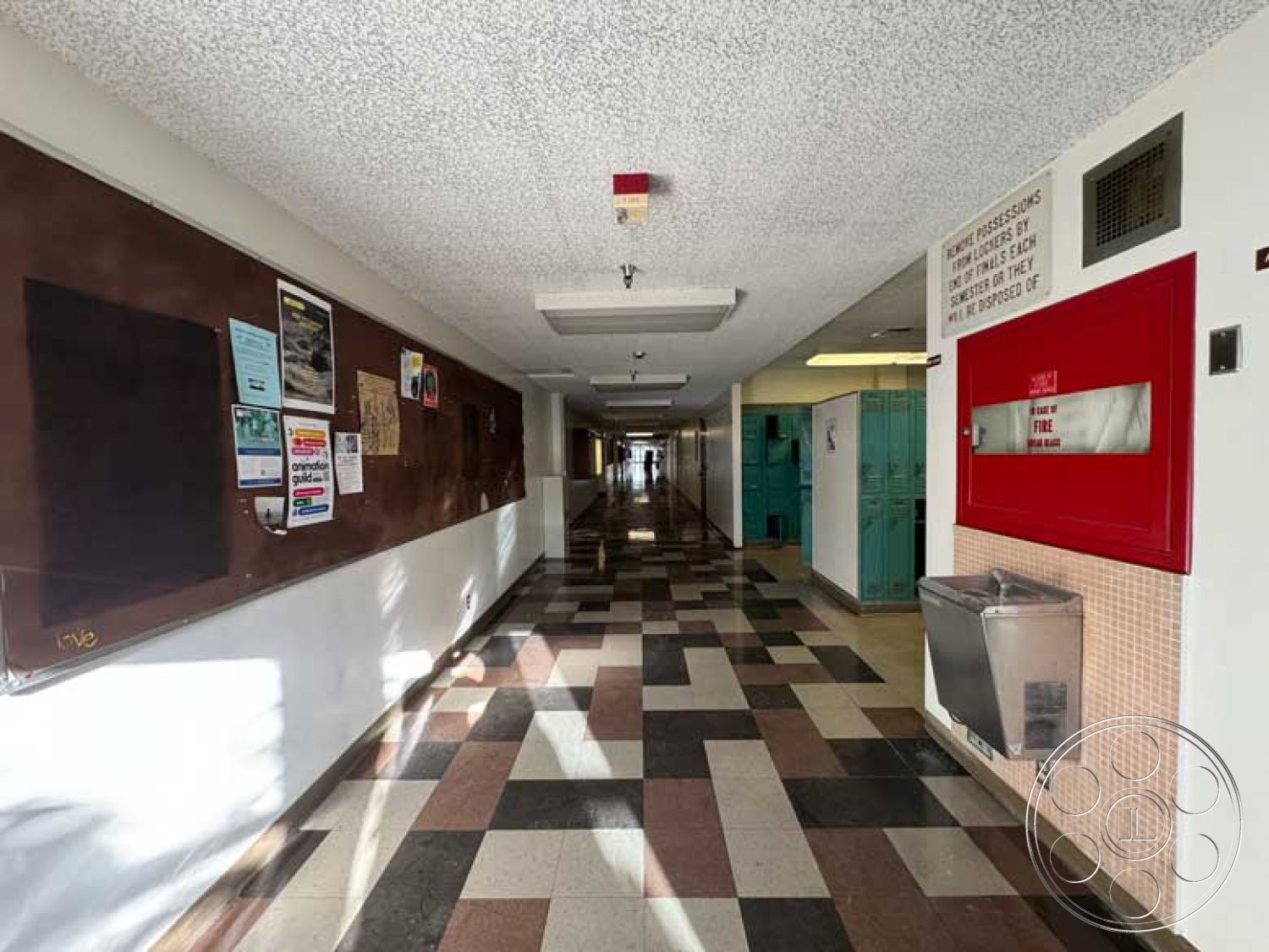 School 3 - painted white walls, polished tile flooring, overhead fluorescent lighting, interior hallway, textured acoustic ceiling panels, emergency exit signage, wide corridor layout, checkerboard pattern tile floors, bulletin board wall treatment, glass drinking fountain, fire alarm box, vintage green lockers