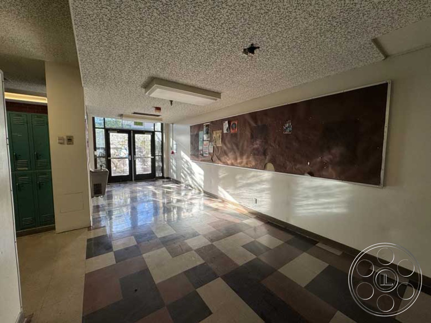 School 3 - textured plaster walls, natural light through windows, large glass entrance doors, exposed ceiling tiles, checkerboard tile flooring, bulletin board wall treatment, lockers with metal finish, corridor layout, interior school hallway, functional communal space