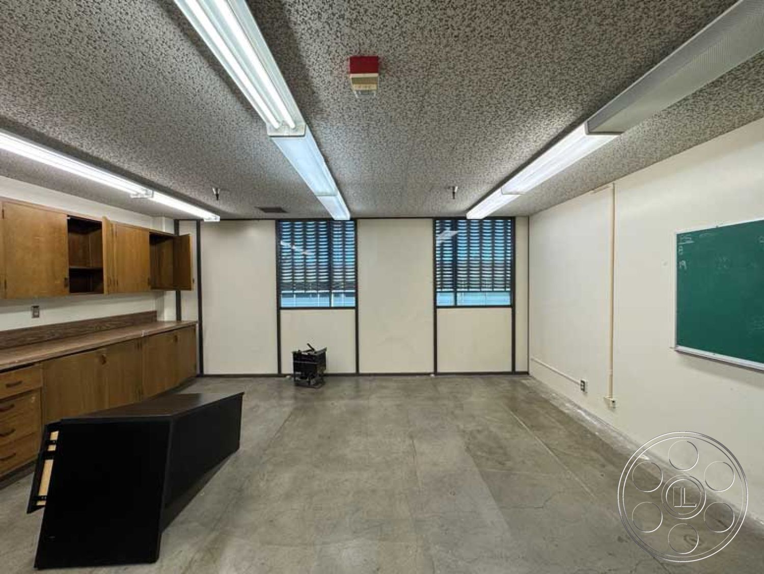 School 3 - neutral color palette, concrete floor, fluorescent lighting, industrial style, functional workspace, natural light from windows, educational environment, open floor plan layout, minimalist desk design, interior classroom space, green chalkboard wall, textured acoustic ceiling, wood cabinetry with laminate finish, steel framed windows with blinds