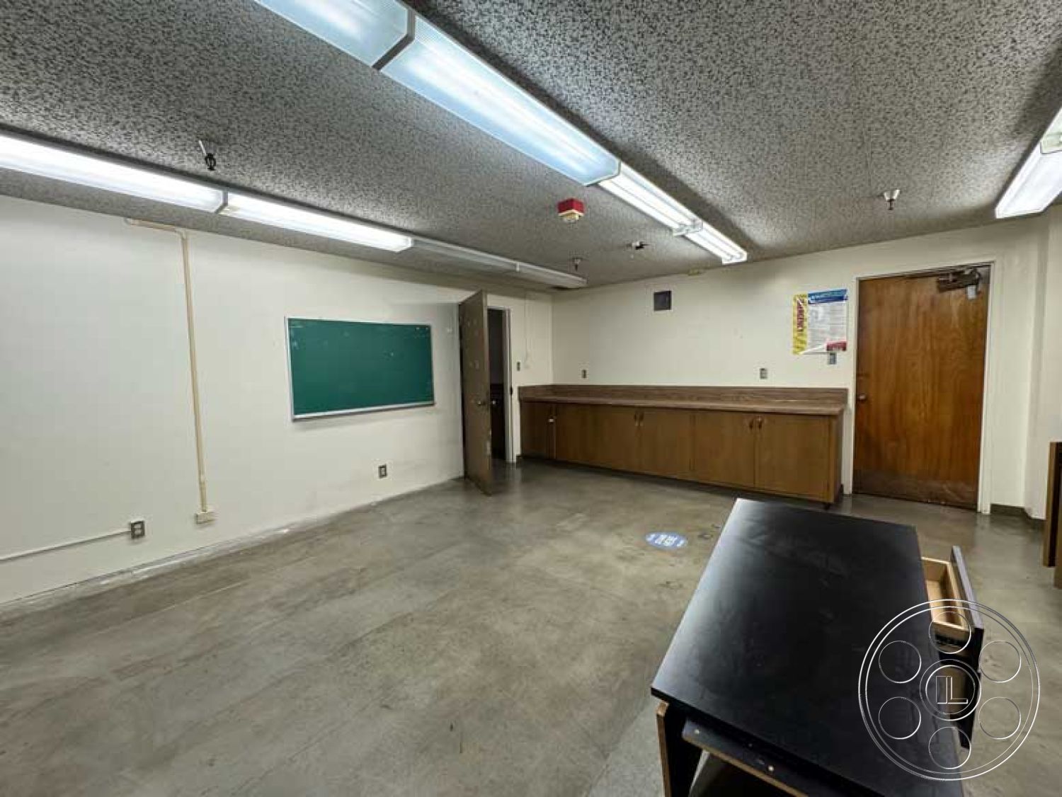 School 3 - chalkboard wall, wooden door frame, open room layout, interior concrete flooring, textured ceiling finish, exposed ceiling tiles, fluorescent ceiling lights, functional classroom setting, laminate cabinetry, industrial style environment