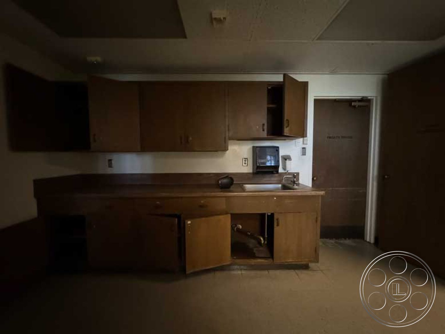 School 3 - neutral color palette, tan tile flooring, modern minimalist style, functional workspace layout, interior kitchen with wood cabinetry, laminate countertop with undermount sink, base cabinets with open doors, ceiling-mounted fluorescent lighting, dark wood paneling on lower walls, white painted upper walls, empty cupboard space, rustic kitchen design, closed door to adjacent room, basic kitchen appliances, single-handle faucet