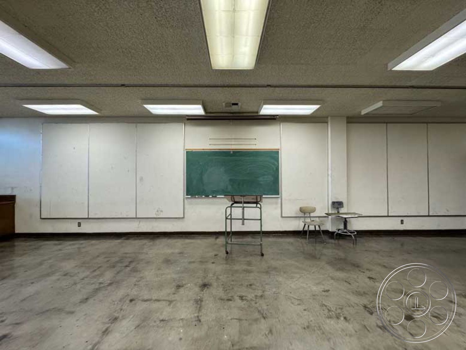 School 3 - neutral color palette, smooth concrete flooring, small round table, school environment, painted drywall walls, textured acoustic ceiling panels, interior classroom space, green chalkboard wall, fluorescent overhead lighting fixtures, industrial metal frame chalkboard, basic wooden cabinetry, stainless steel desk surface, folding chair with metal legs, empty classroom setting