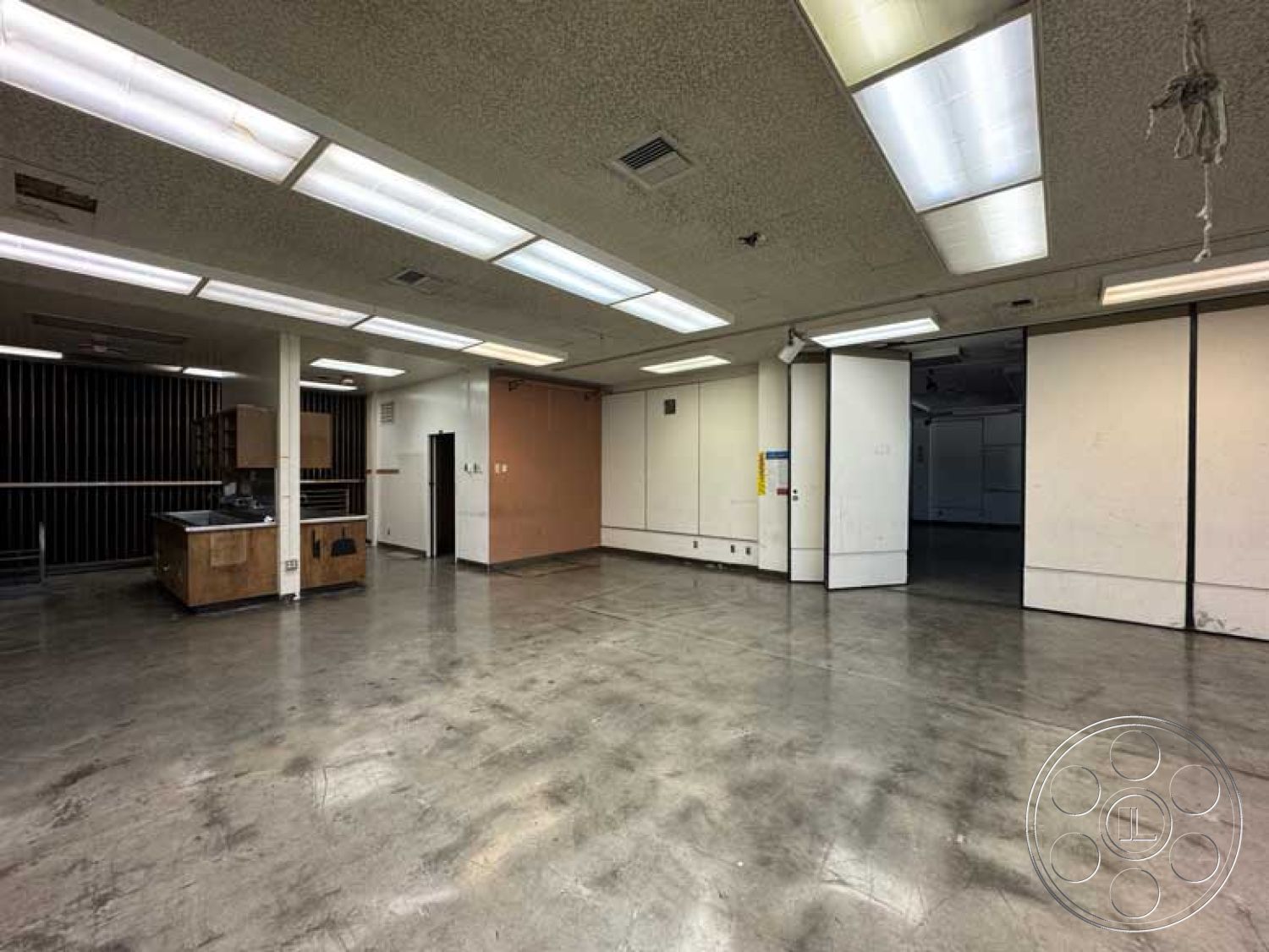 School 3 - urban setting, minimalist design details, glass door entrance, exposed concrete flooring, open layout interior, commercial space interior, industrial-style lighting, drop ceiling with fluorescent lights, drywall partition walls, utility sink area, floating shelves in cabinetry, neutral color scheme walls, wood laminate kitchen cabinets, vinyl composite tile floor