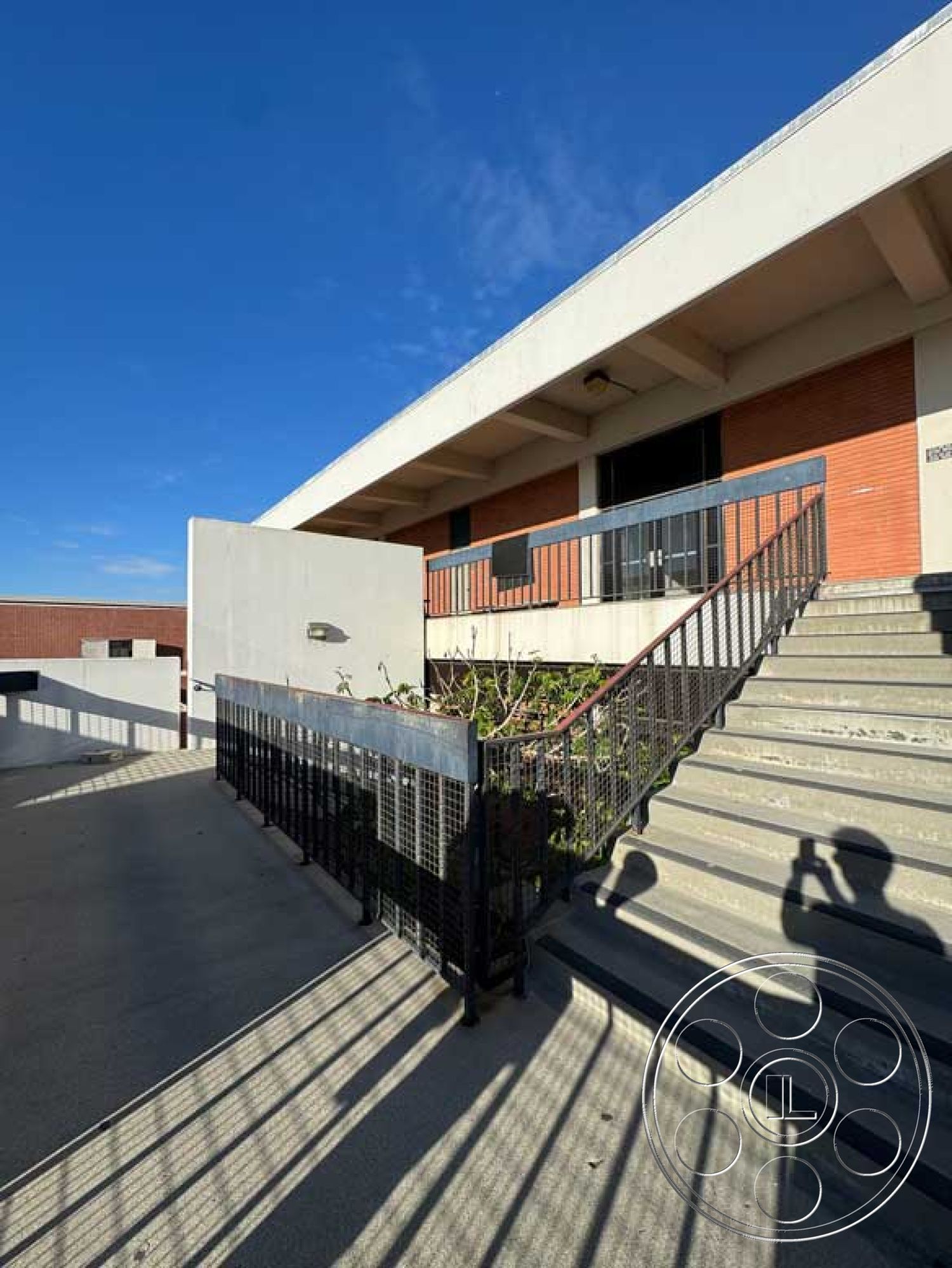 School 3 - industrial style, urban setting, exposed brick wall, textured wall treatment, modern architectural design, open sky view, flat roof architecture, exterior concrete stairs, landscaped planter area, horizontal lines in design, steel railing with mesh accents, concrete landing, shade from overhanging roof, shadow play on concrete surface