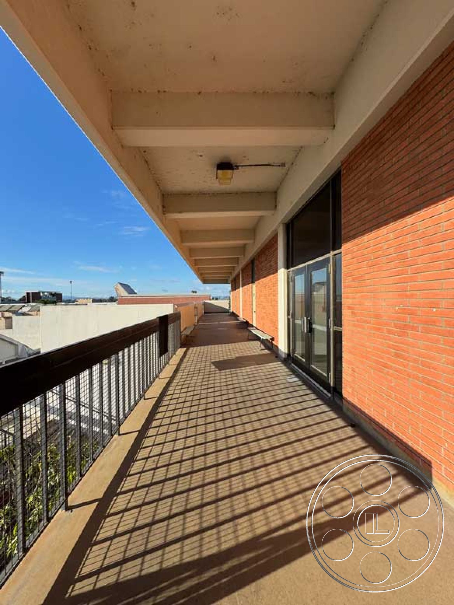 School 3 - accessible outdoor area, flat roof structure, floor-to-ceiling glass doors, industrial architectural style, urban landscape view, exposed brick exterior walls, sunny weather environment, natural light source, concrete balcony flooring, metal railing with vertical bars, modern commercial building design, open-air walkway, interior/exterior transitional space, concrete support beams, rectangular balcony shape