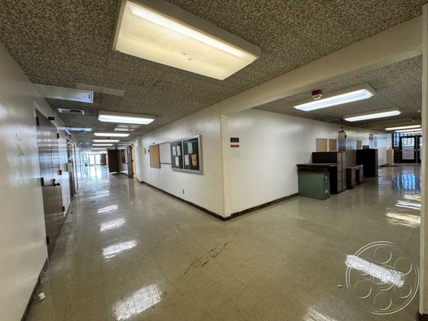 School 3 - smooth wall finishes, commercial hallway design, interior corridor with vinyl tile flooring, exposed acoustical ceiling tiles, white painted drywall walls, flush-mounted fluorescent ceiling lights, metal door frames, black bulletin board with mounted papers, polished concrete baseboards, public building corridor layout