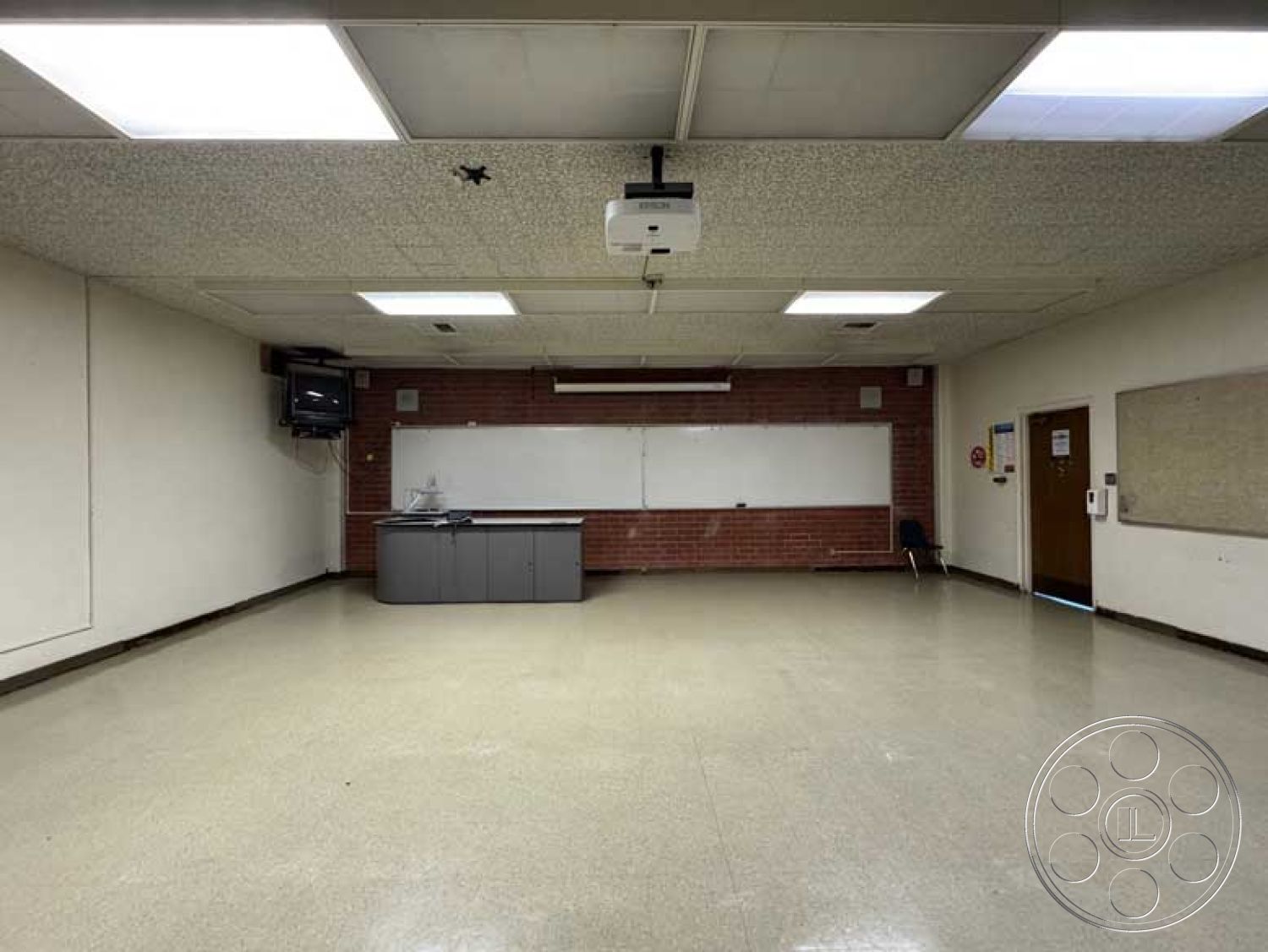 School 3 - acoustic ceiling tiles, exposed brick accent wall, fluorescent lighting fixtures, whiteboard wall, built-in storage cabinet, interior classroom, overhead projector, smooth linoleum flooring, dark wood paneling trim, neutral color painted walls, single desk setup, classroom environment
