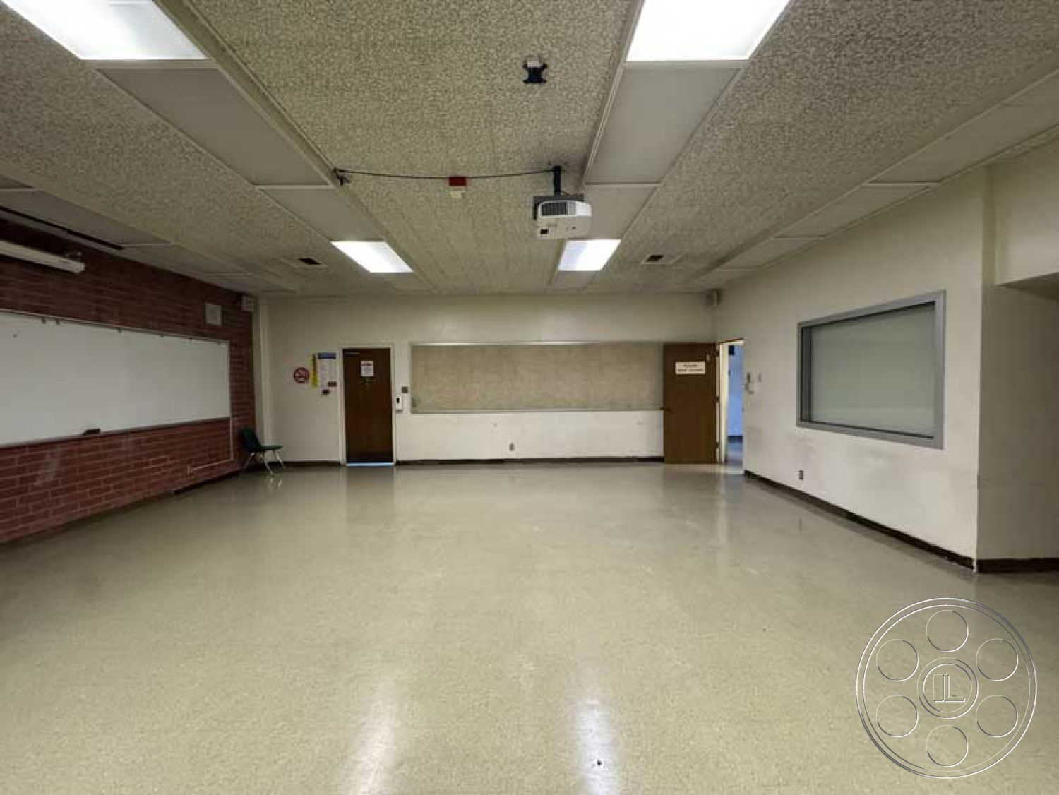 School 3 - vinyl flooring, brown wooden doors, acoustic ceiling tiles, white painted walls, exposed brick accent wall, interior classroom space, flush-mounted fluorescent lights, window with glass partition, projector mount on ceiling, classroom wall with whiteboard