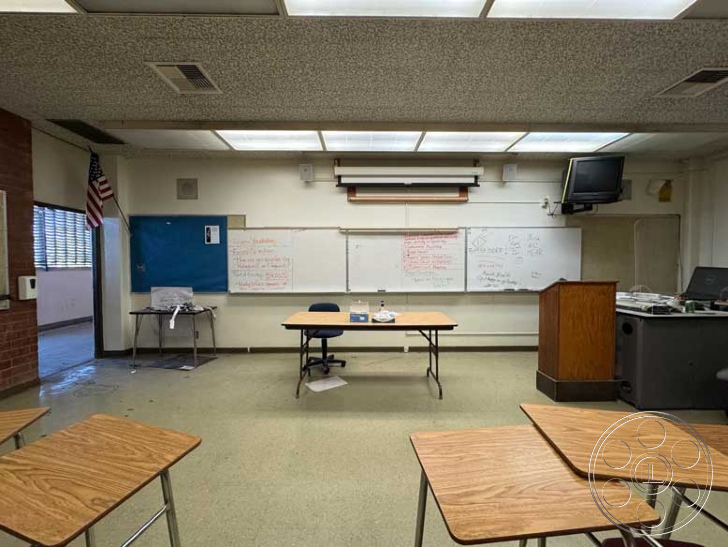 School 3 - brick accent wall, vinyl flooring, textured acoustic ceiling panels, fluorescent ceiling lights, whiteboard wall, overhead projector, interior classroom space, open classroom layout, wooden classroom desks, steel chair legs, cork bulletin board, blue chalkboard wall, teachers' podium, projector screen attachment, windowed classroom, American flag display, traditional educational space, functional learning environment, school setting