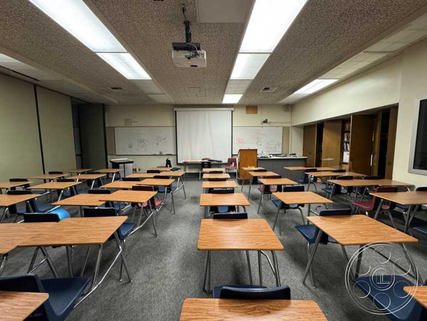 School 3 - neutral wall colors, projector screen, acoustic tile ceiling, whiteboard wall, drop ceiling with fluorescent lighting, double door entrance, interior classroom setting, black plastic stacking chairs, vinyl composition tile flooring, laminate teacher's desk, wood grain school desks, wall-mounted chalkboards, library bookshelf alcove, centralized classroom layout